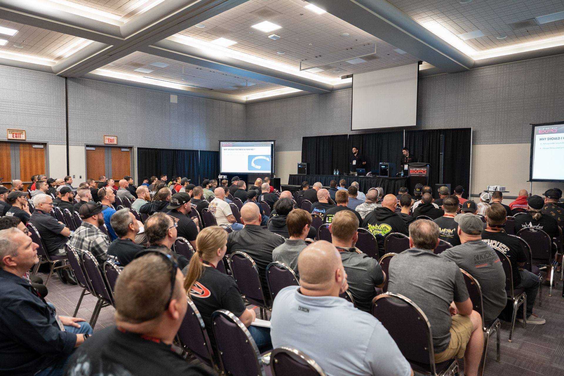 Audience seated in a large conference room, listening to a presentation on a stage with screens.