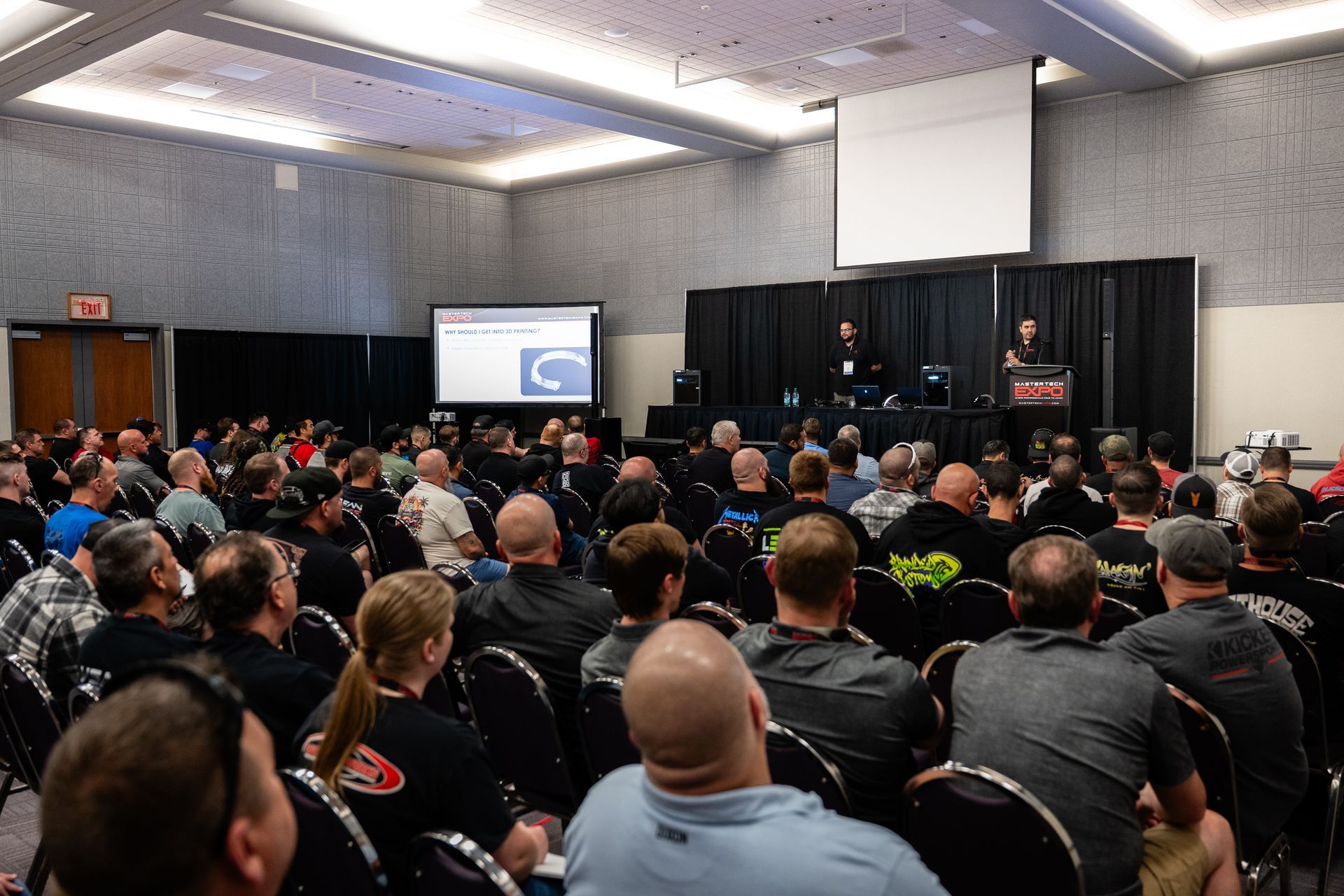 Audience at a conference listens to presenters on stage. Large room, screen, and black curtain backdrop.
