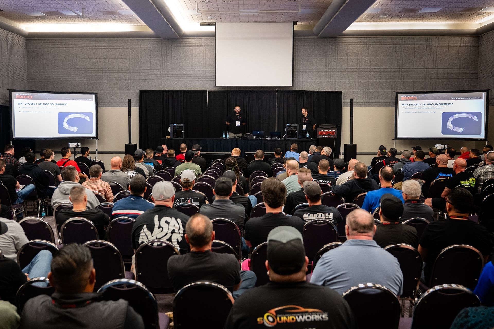 Audience at a conference listens to presenters on stage, three screens show a diagram.