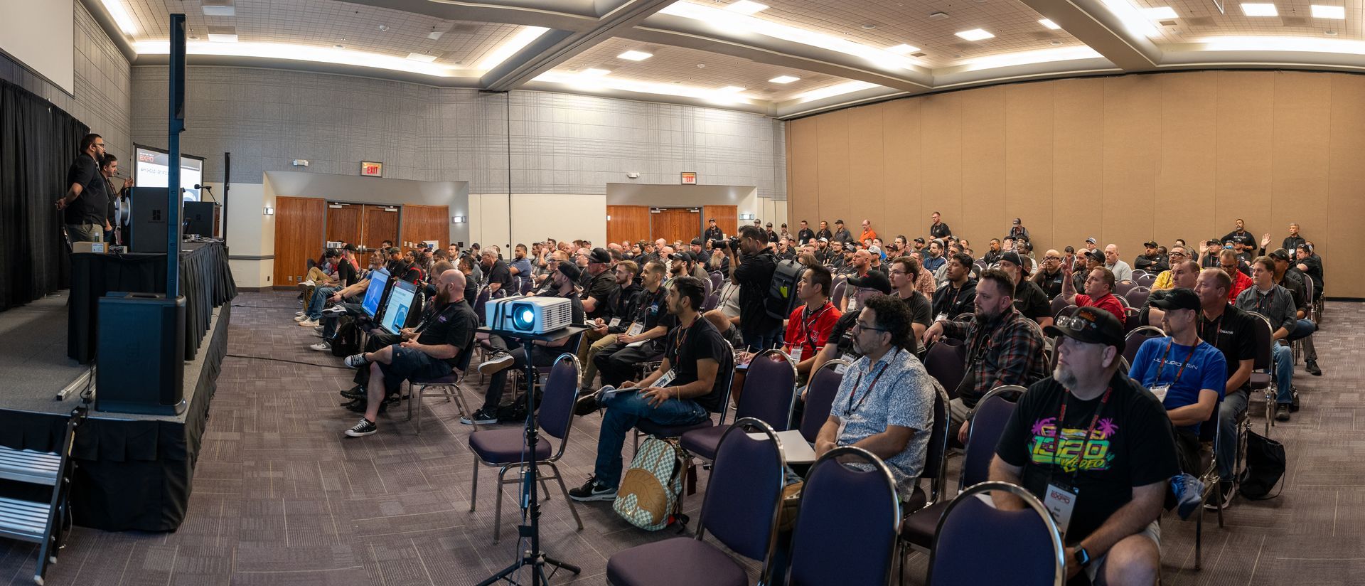 A large conference room with a presentation in progress. A projector displays an image, many people are seated and listening.
