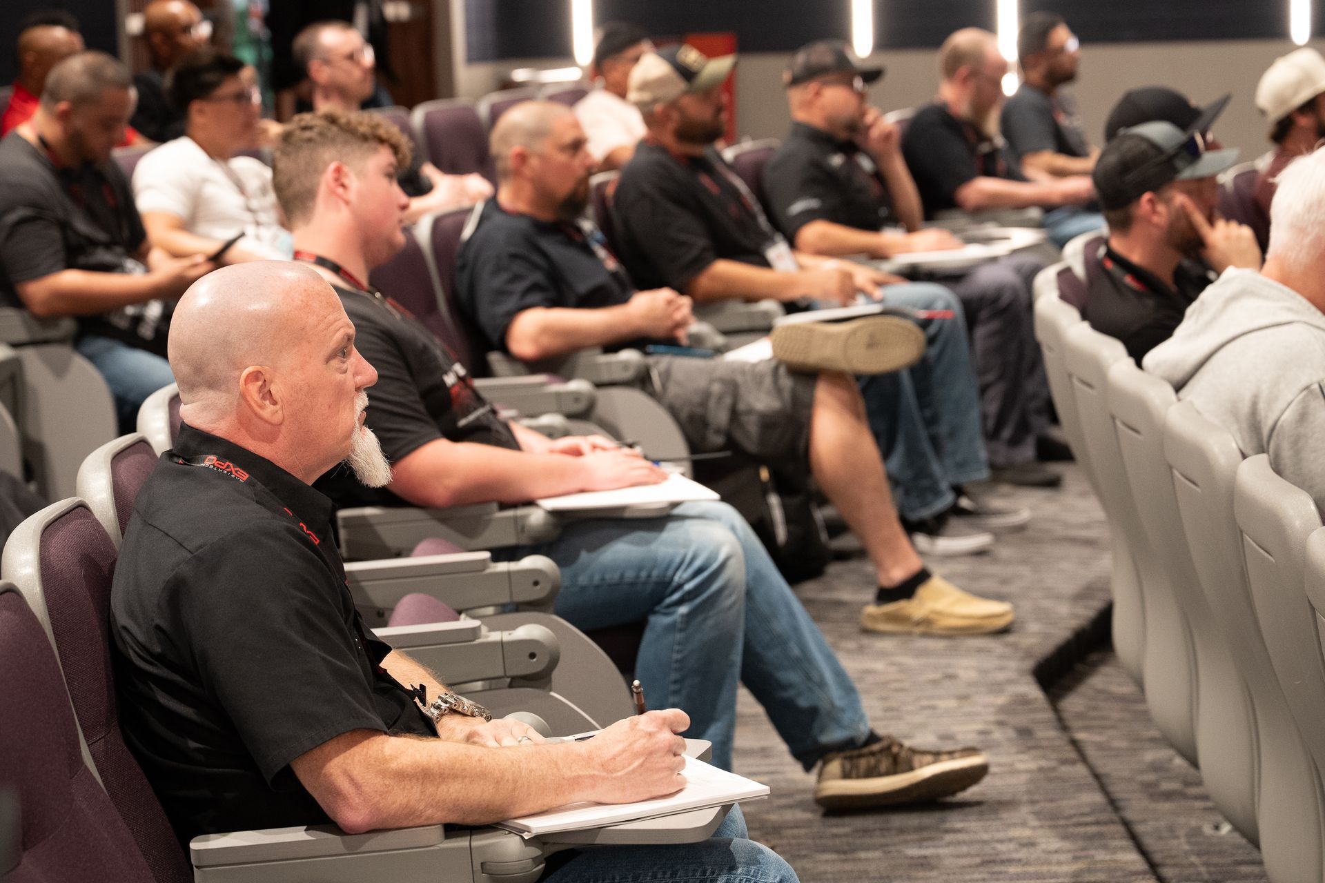 People seated in rows, listening to a presentation; some take notes. Dim lighting, neutral tones.