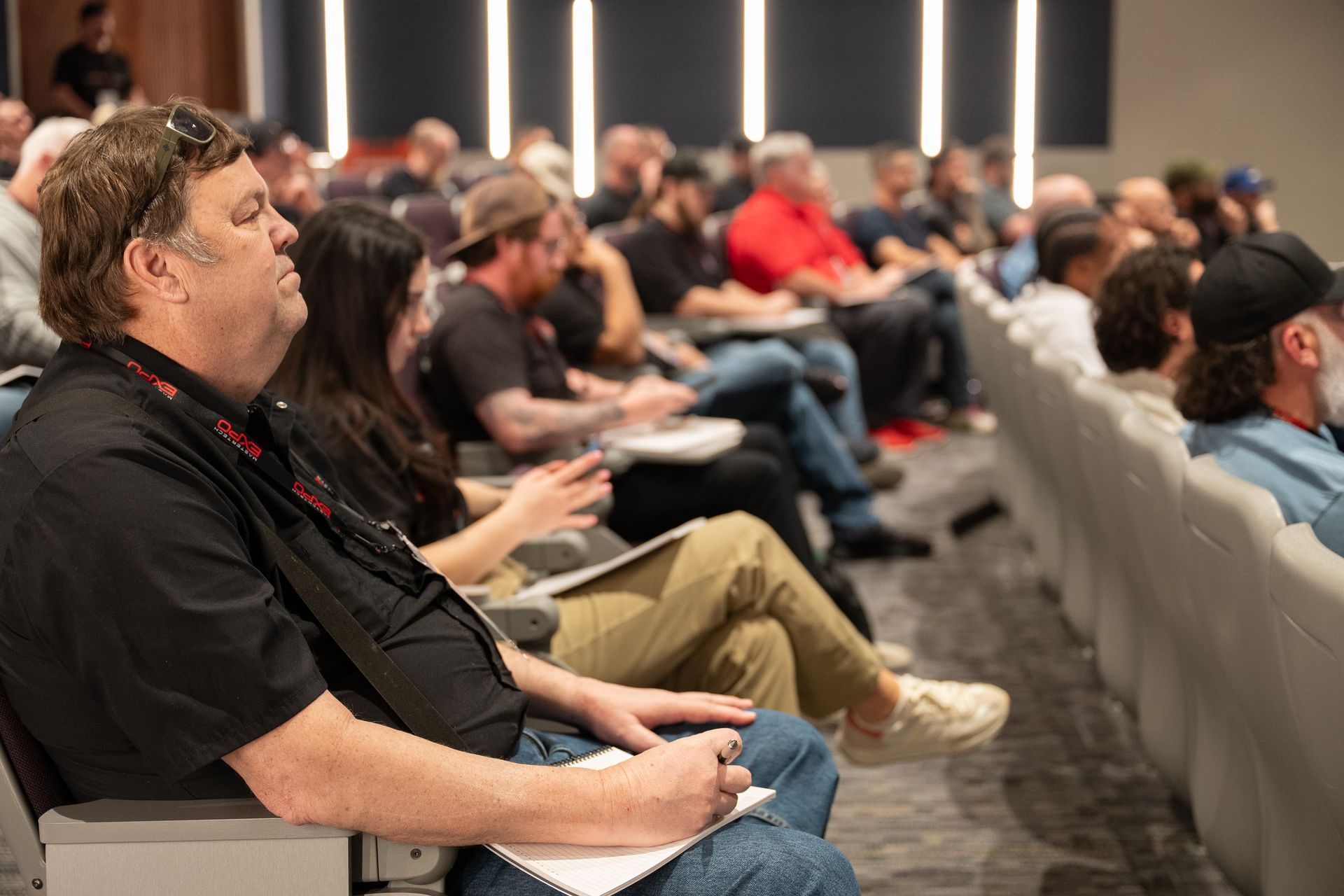 Audience in a conference room; people seated, taking notes; a man in a black shirt looks toward the presenter.