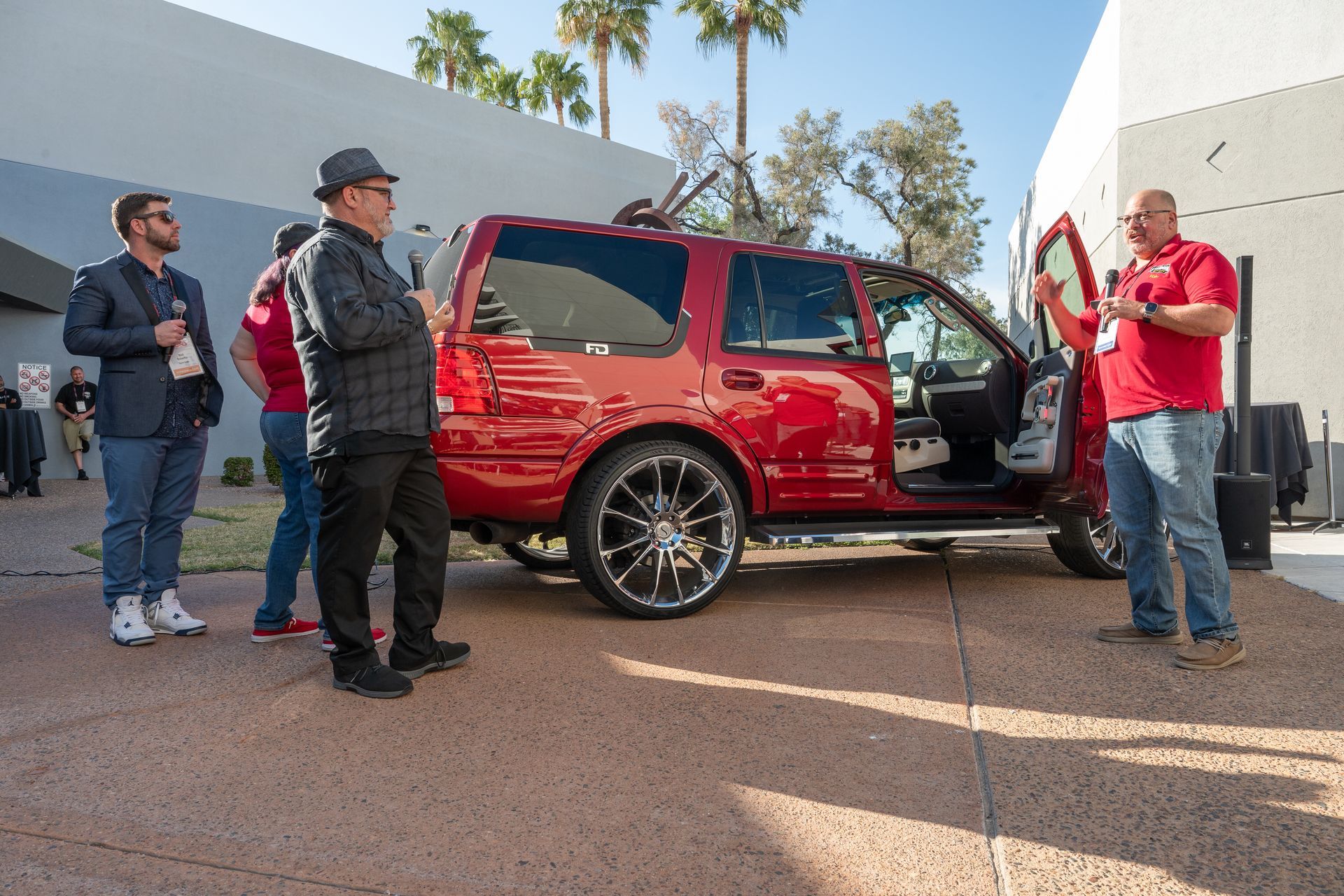 People look at red SUV with open door; person in red shirt gestures, sunny outdoor setting.