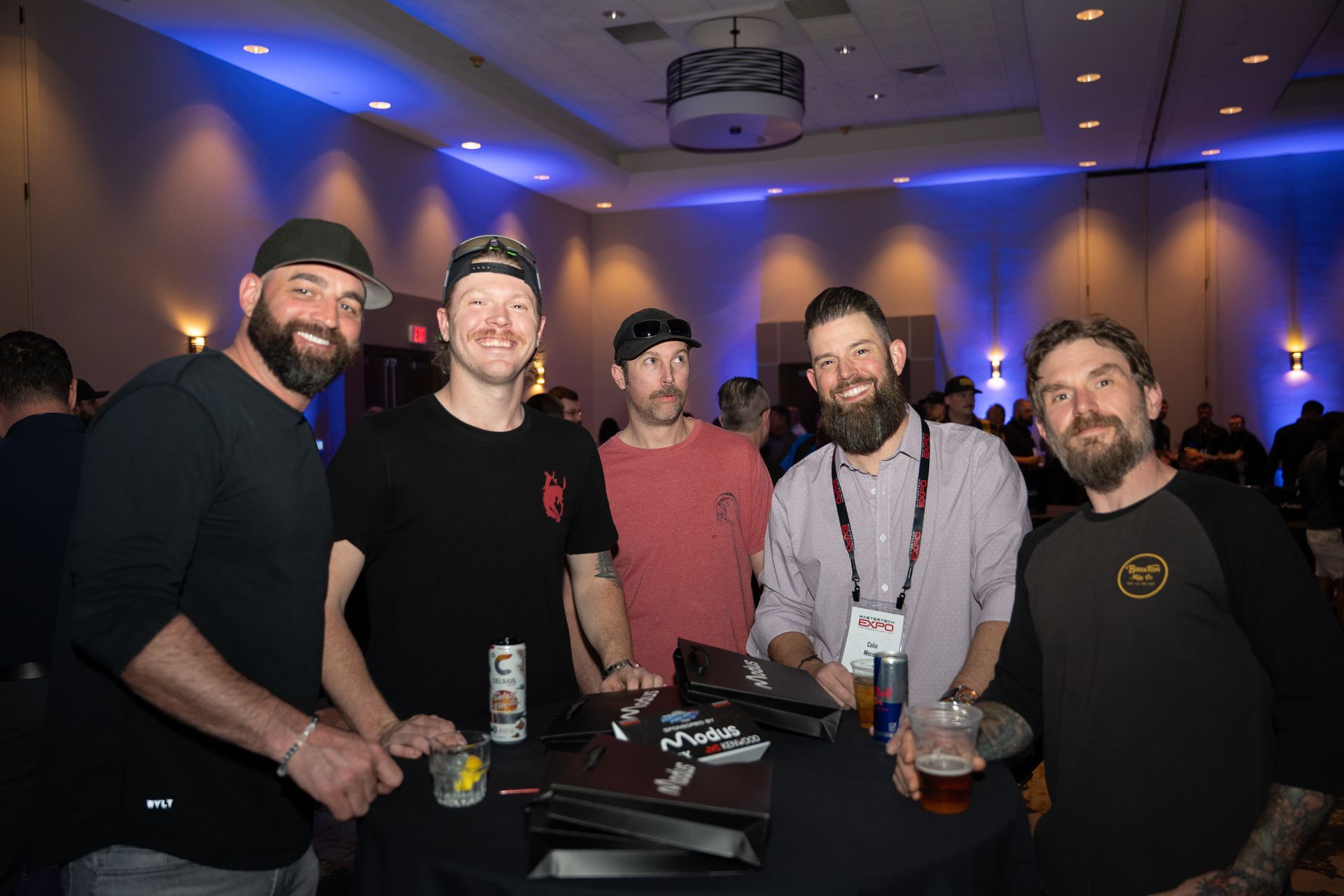 Five men with beards smile at the camera at an event, gathered around a table with drinks, in a room with blue and yellow lighting.