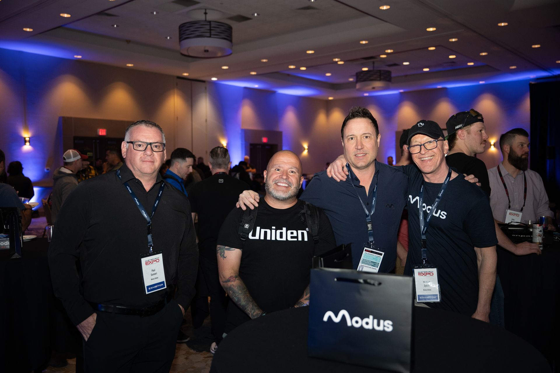 Four men posing at a Modus branded table, lit with blue and white lights in a room.