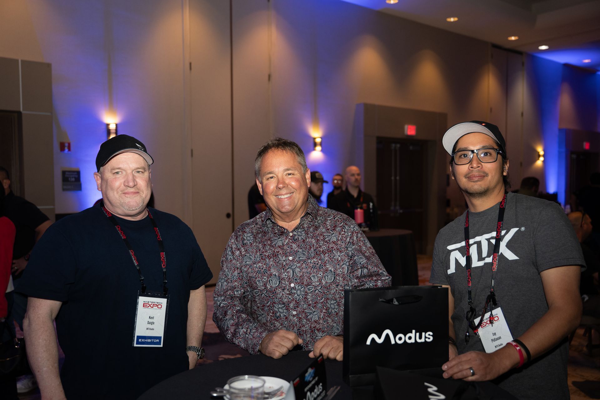 Three men at an event pose by a table. A 