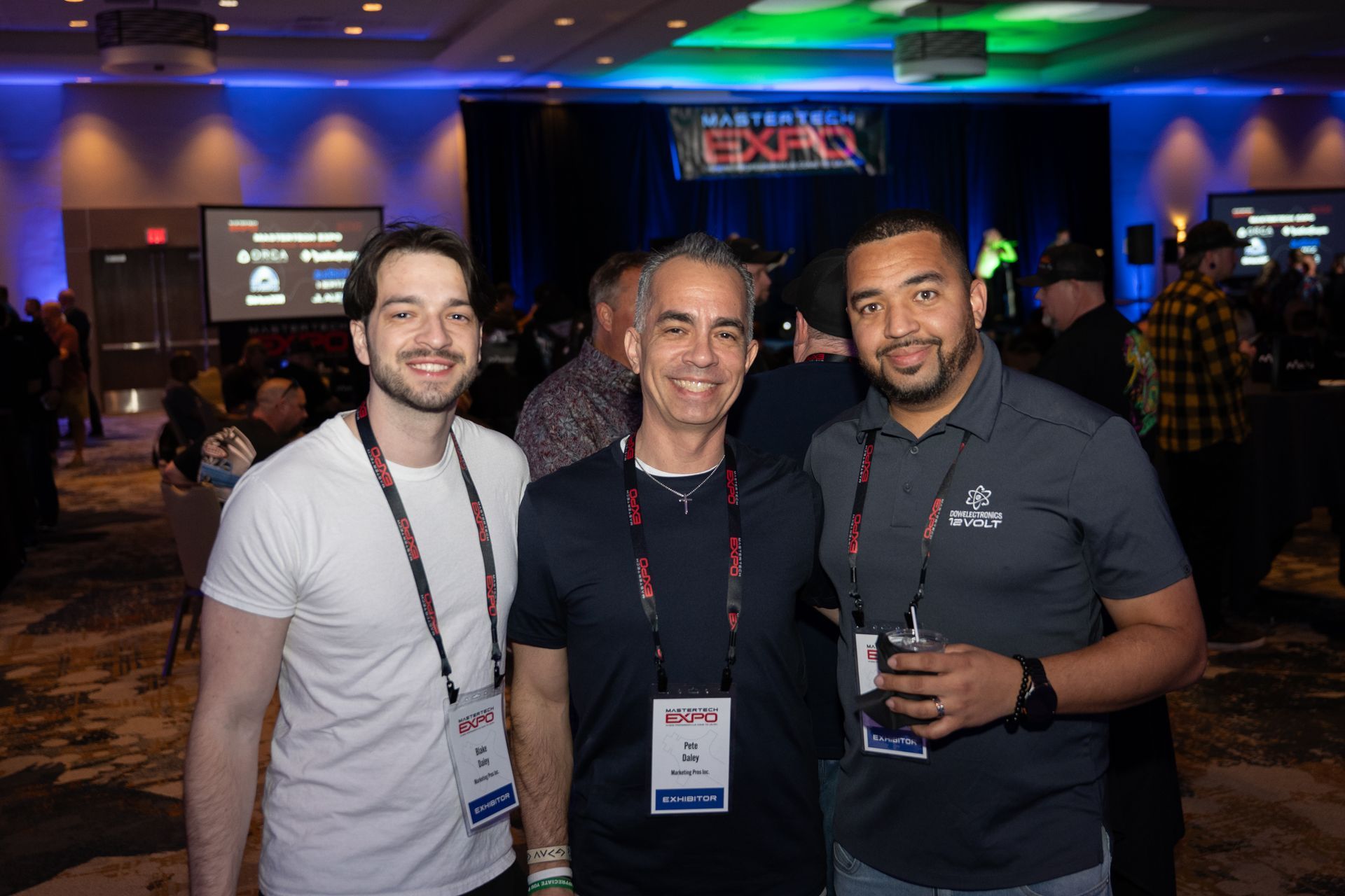 Three men at a conference pose for photo. One in white shirt, one in navy polo, and one in a gray shirt, all wearing lanyards, smiling.