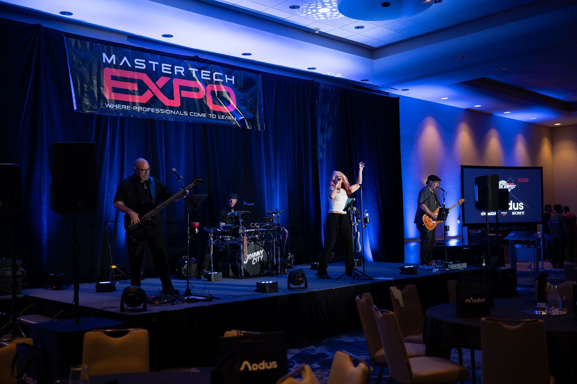 Band performing on stage at the Mastertech Expo. Dark clothing, blue lighting, banner backdrop.