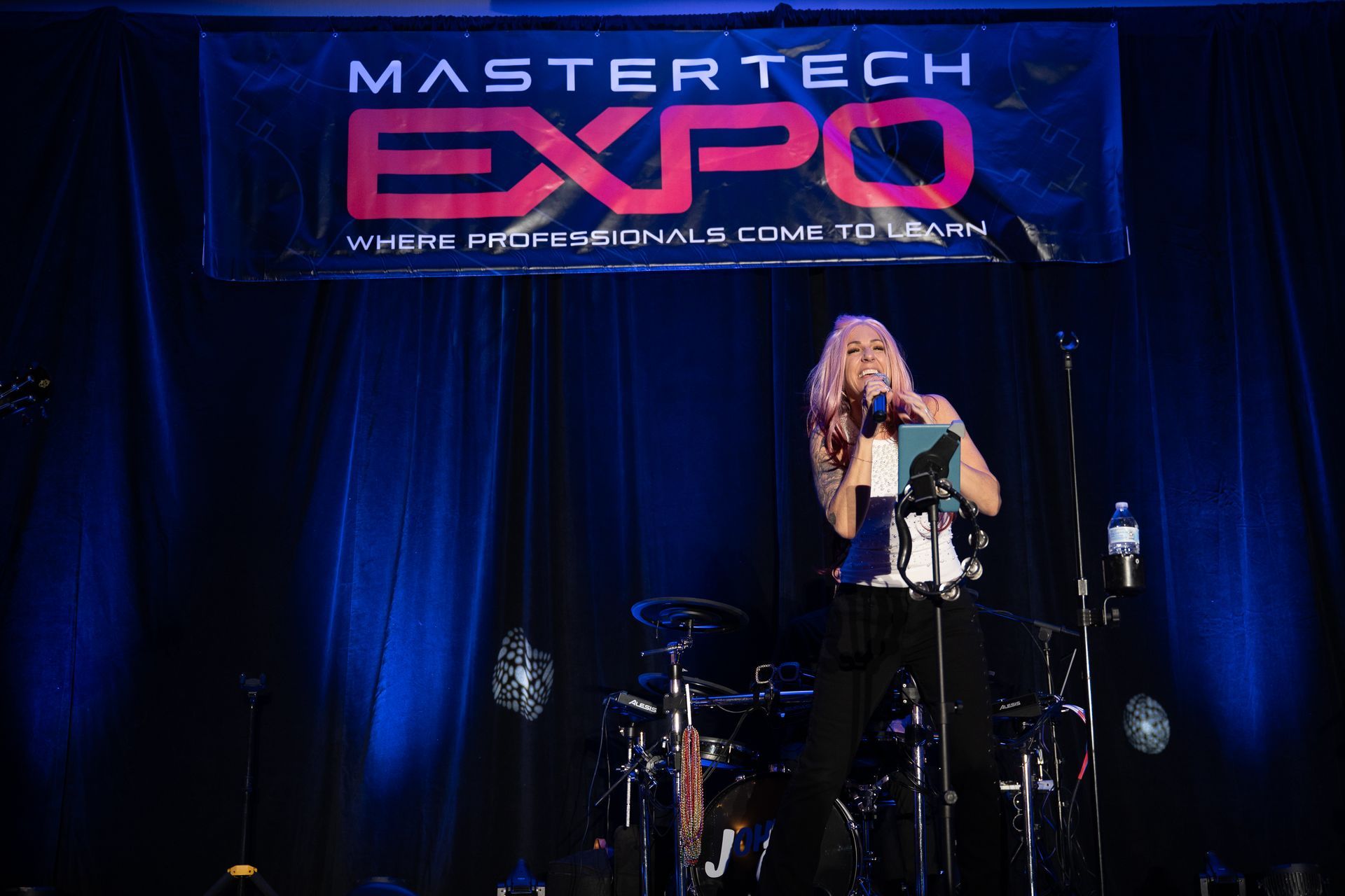 Woman on stage performing at Mastertech Expo; blue backdrop, drums, microphone.
