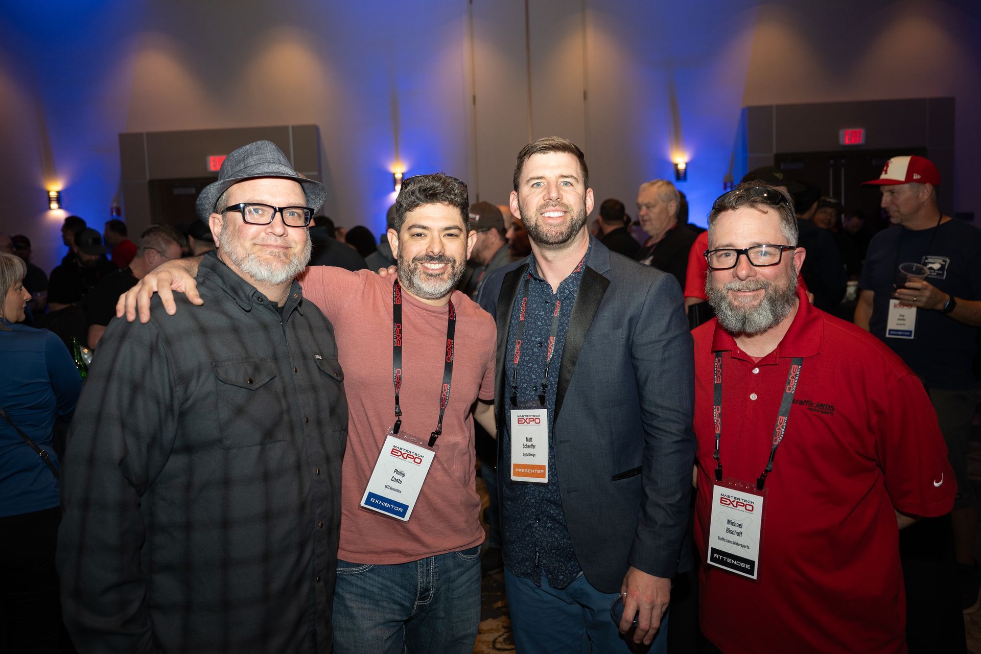 Four men pose together at an event. They wear lanyards and smile. The room has blue lighting.