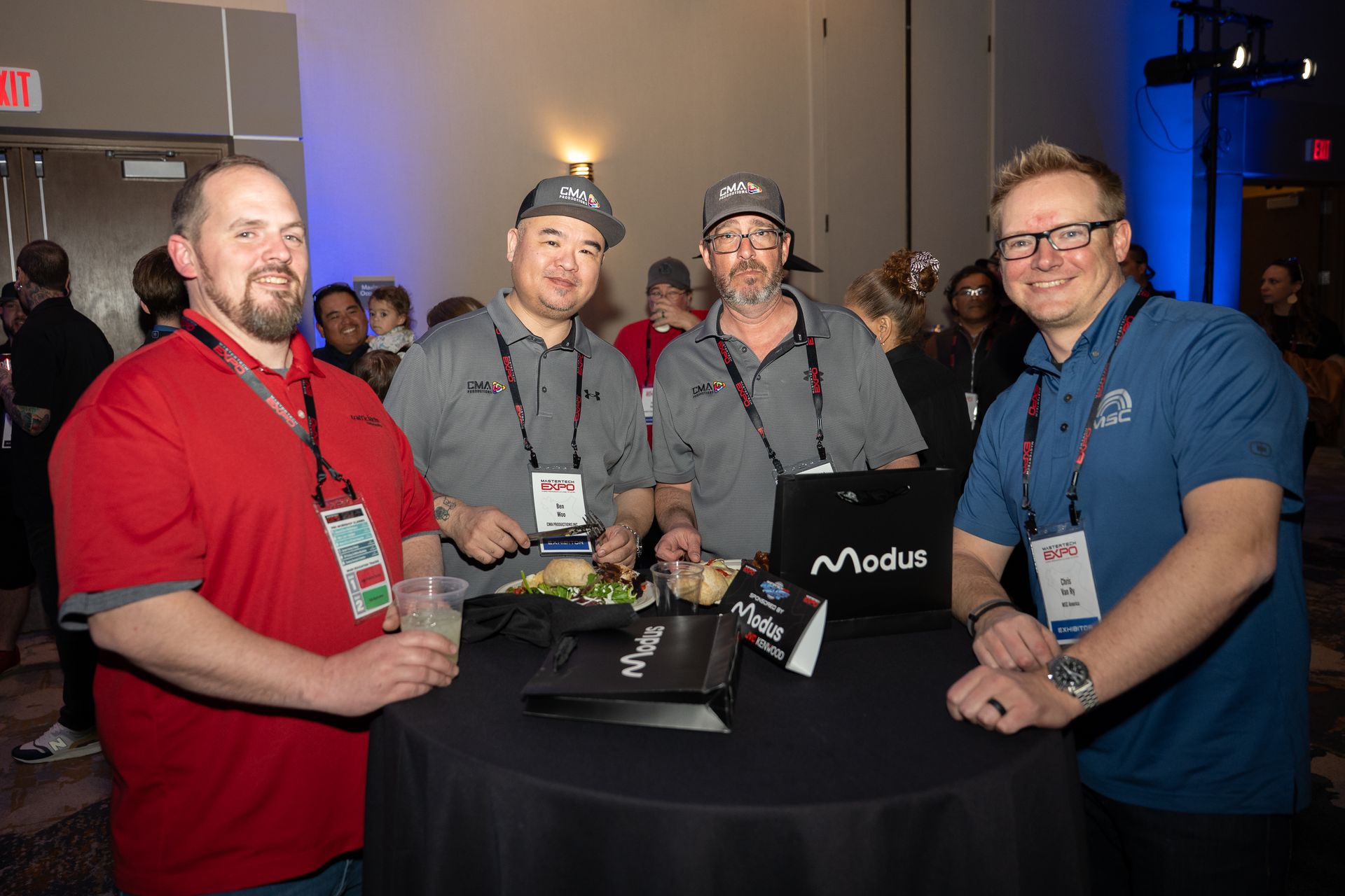 Four men at a table, some holding food and drinks, wearing lanyards and hats. Lit room, event setting.