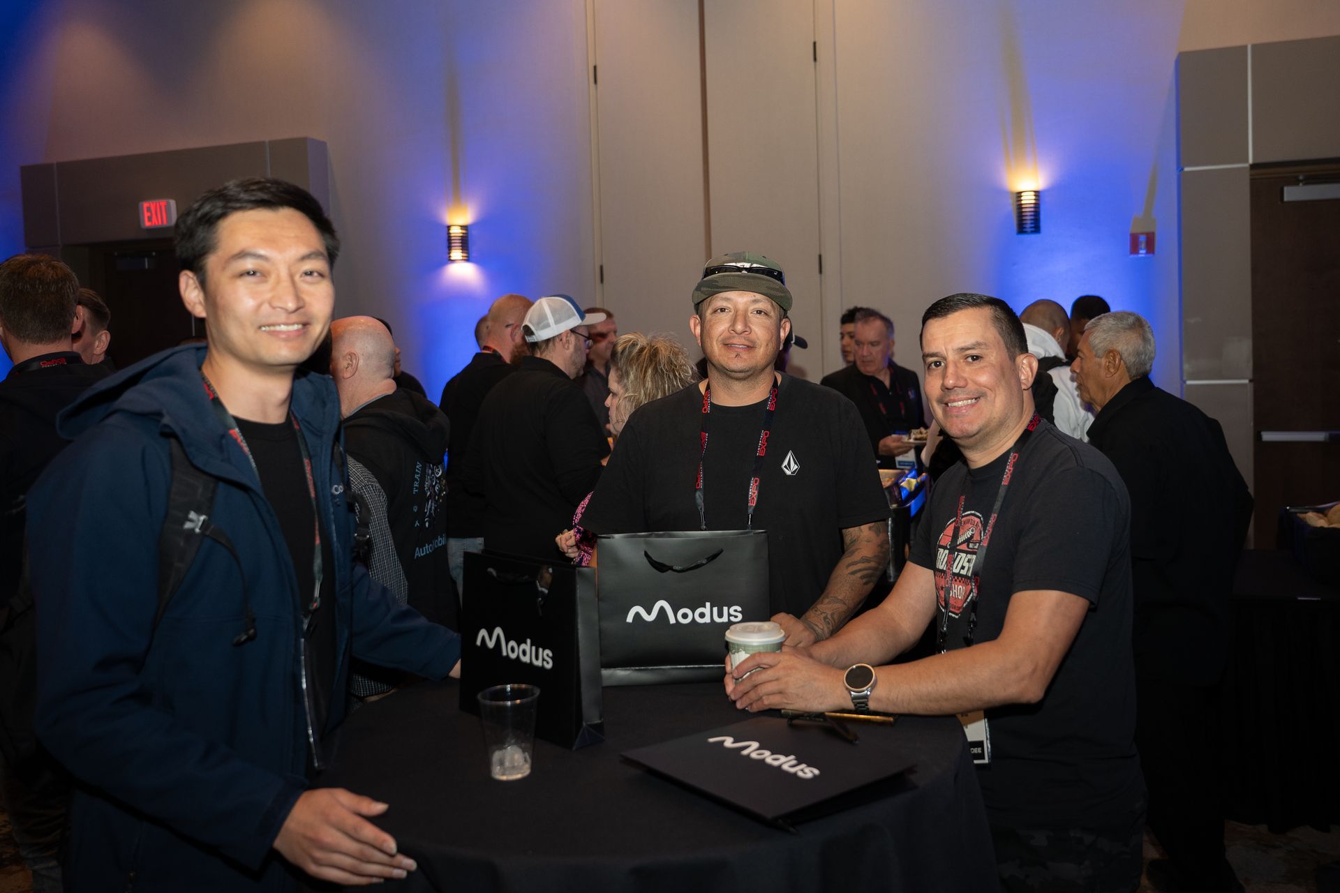 Three men at an event with others in the background, one holding a gift bag, one with a coffee cup.
