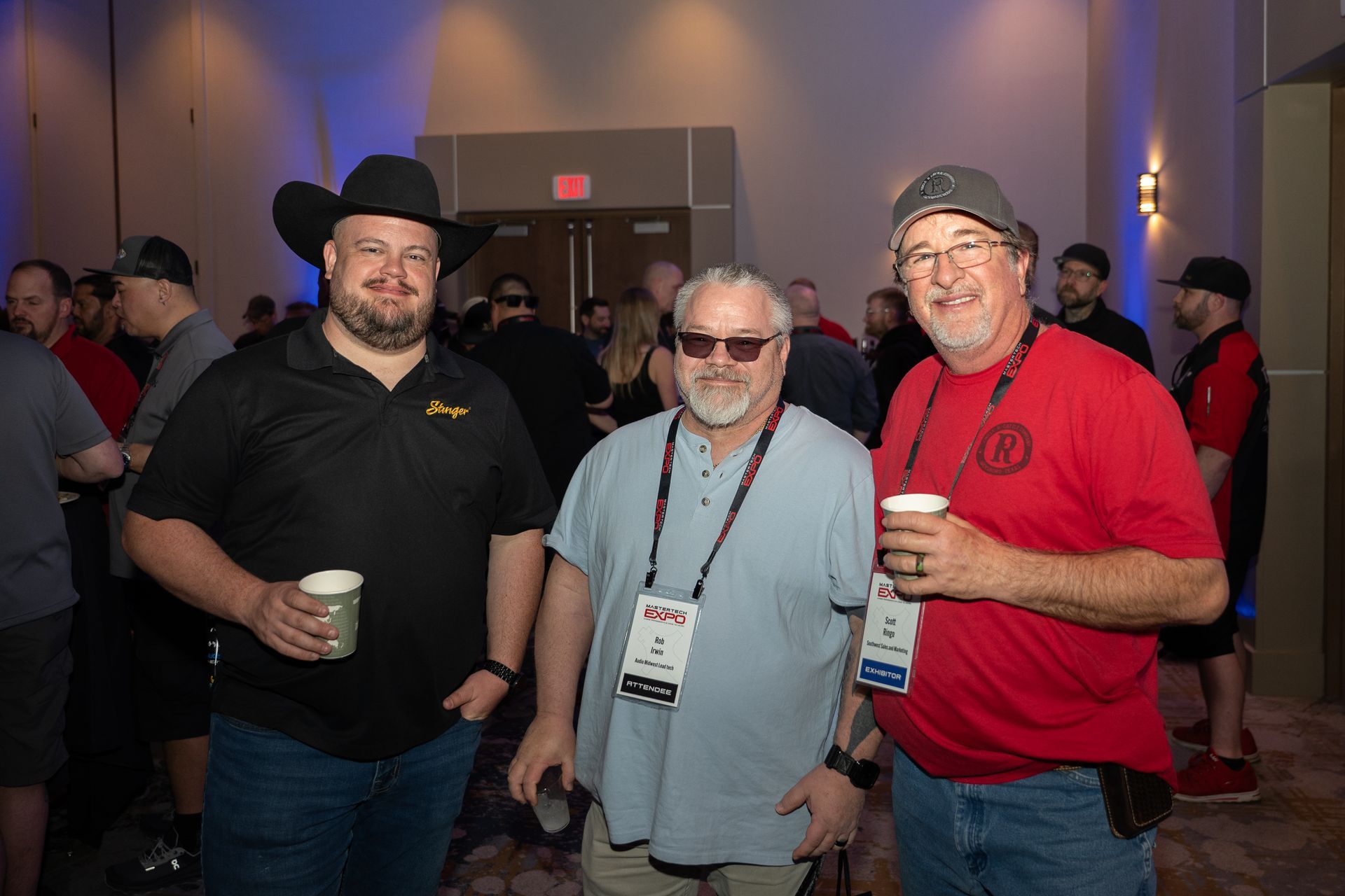 Three men at event, holding drinks, smiling. One wears cowboy hat, another sunglasses, and the third a cap and red shirt.