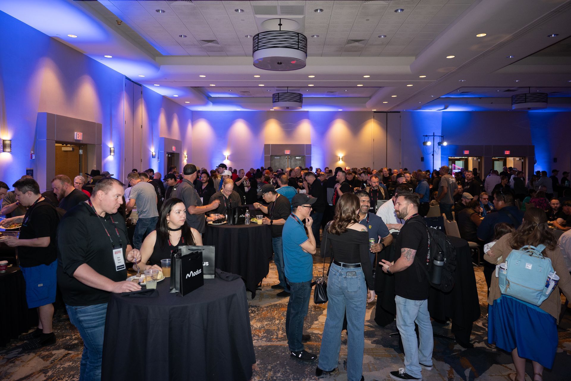 Large crowded event in a brightly lit ballroom with people gathered around tables, socializing. Blue and yellow lighting.