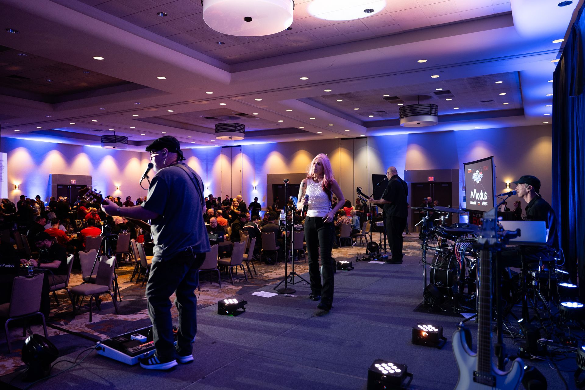 Band performing on stage at an event, with audience in background. Blue and white lighting.