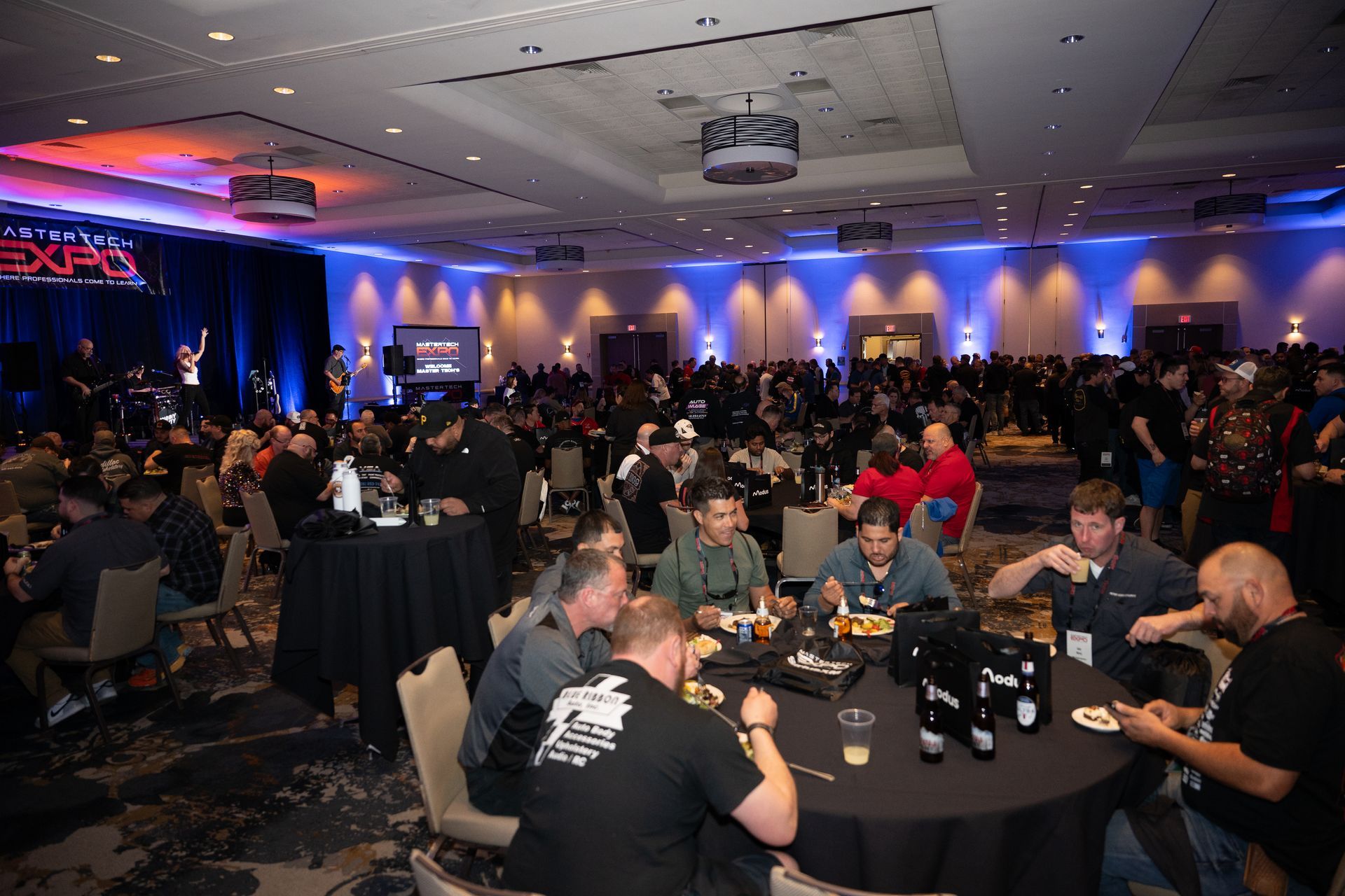 Large event in a ballroom with many attendees at tables; stage at the front with blue and red lighting.