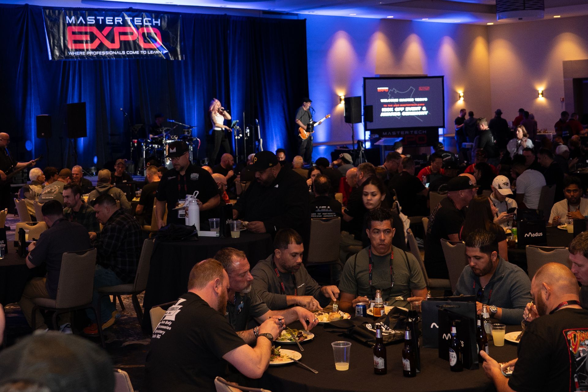 A crowded expo hall with a live band on stage. People at tables are eating and socializing.