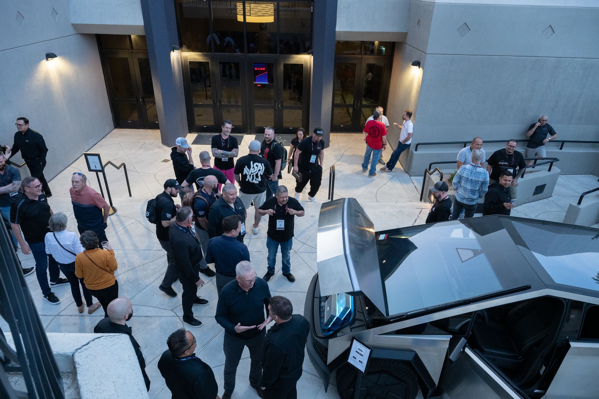 People gather outside a building, near a silver truck. Some wear black, others casual attire. Evening setting.