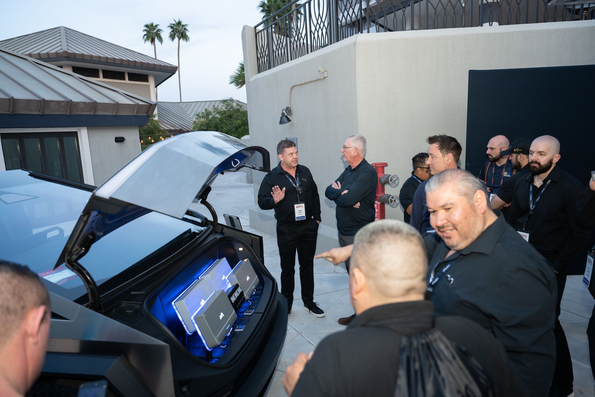 A man explains a car's audio system to a group outdoors. Car's trunk is open, revealing speakers.
