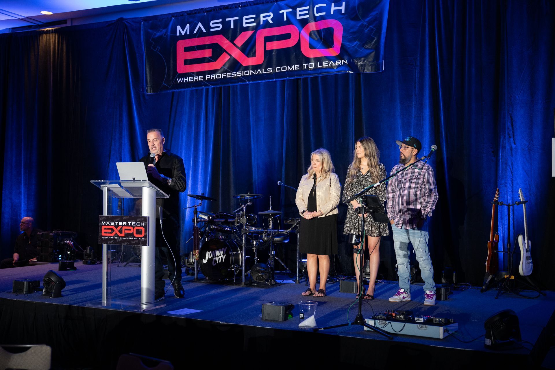 A man speaks at a podium. Three others stand behind him on a stage with a 