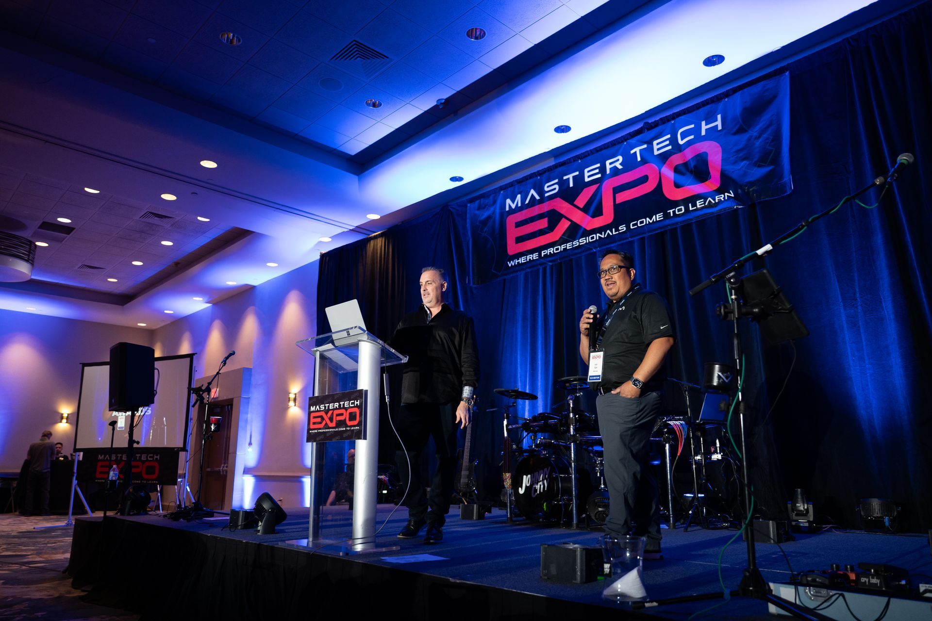 Two men on stage at the Mastertech Expo. One speaks, the other holds a microphone. Backdrop banner and stage lights.