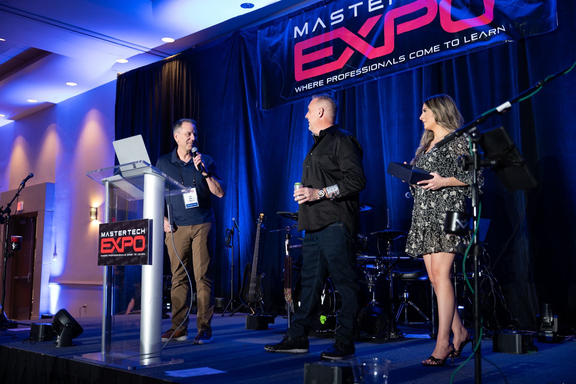 Three people on stage at an expo. Man speaking into microphone, two others listen. Stage backdrop with event logo.