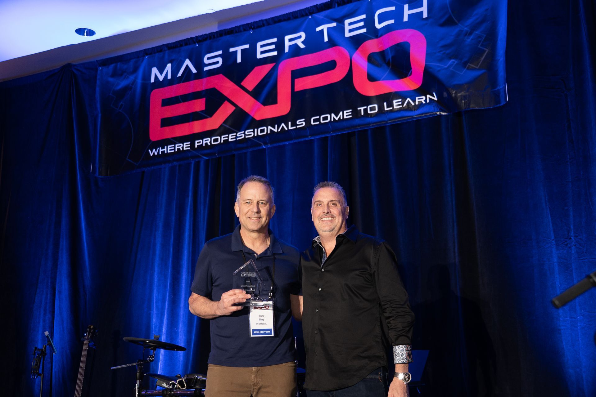 Two men on stage holding an award at the Mastertech Expo. A banner is in the background.