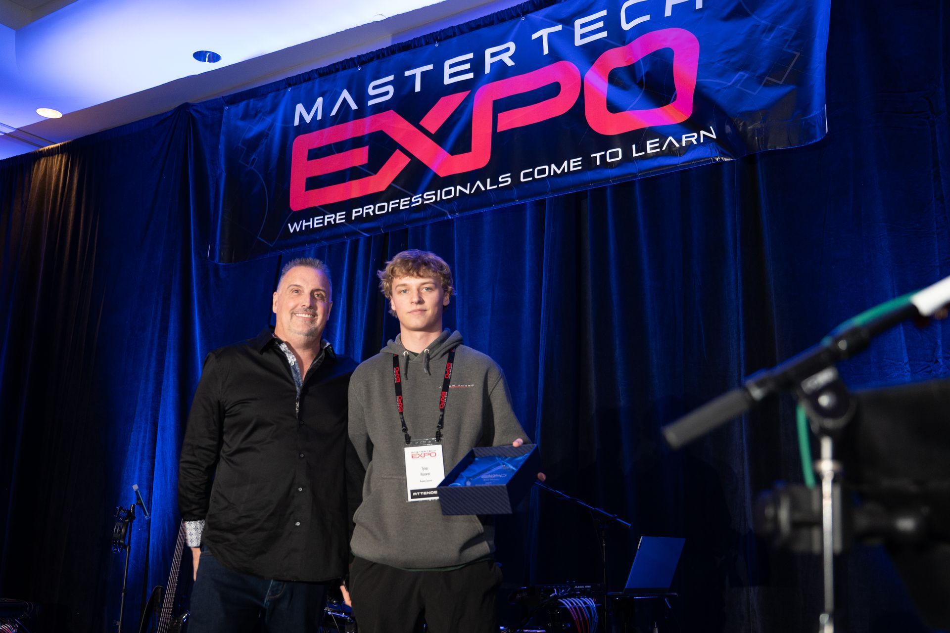 Two men at a Mastertech Expo event. One presents an award to a young man holding a plaque, in front of a blue backdrop.
