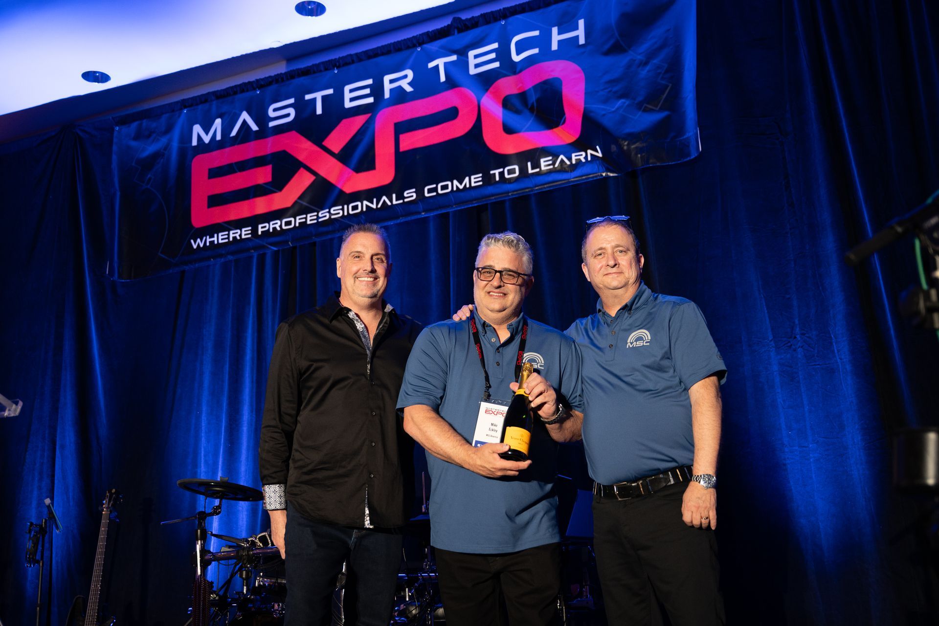 Three men holding a bottle on stage at the Mastertech Expo. Banner in the background.