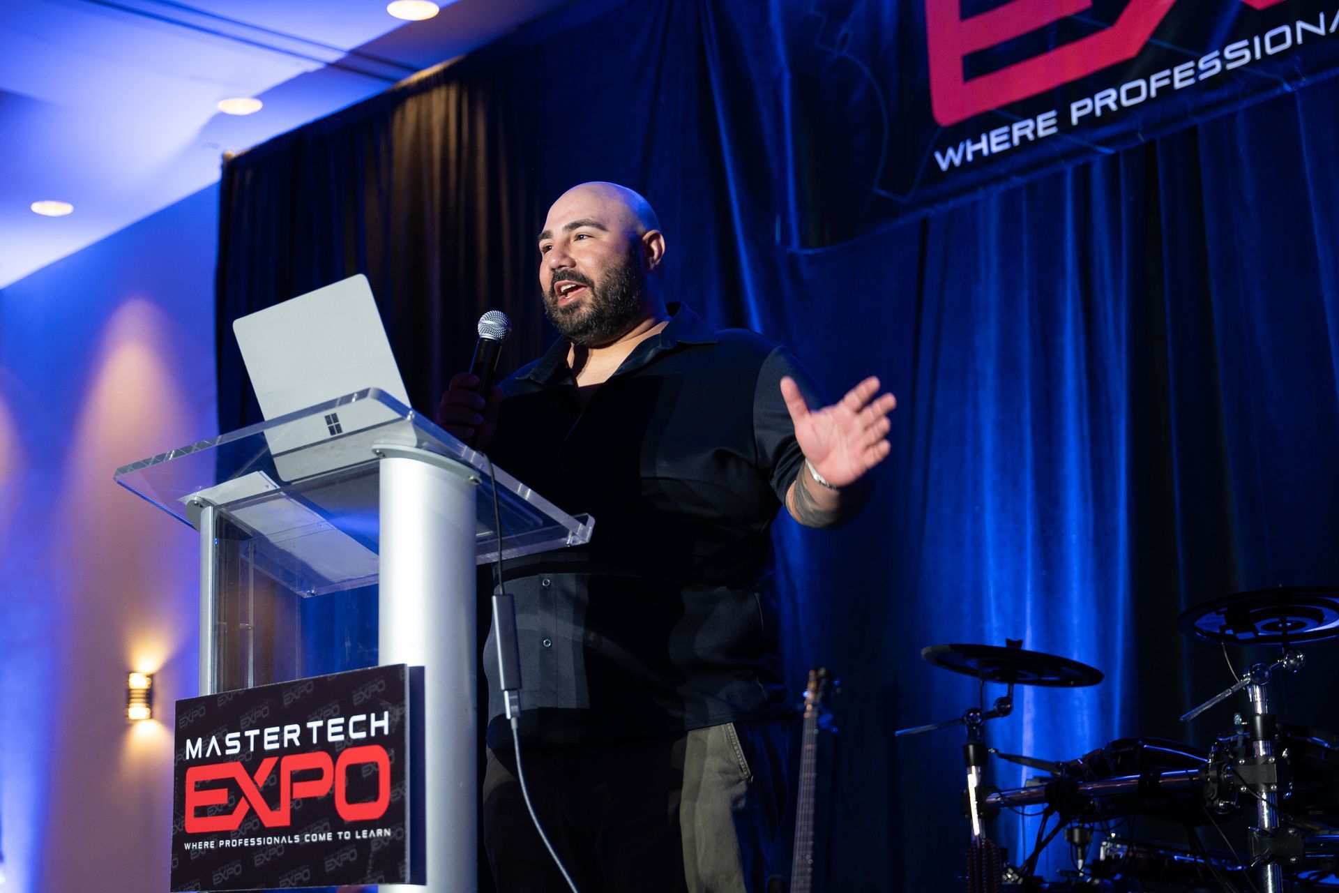Man speaking at a podium during Master Tech Expo. He gestures with hands, laptop on stand, with blue background.