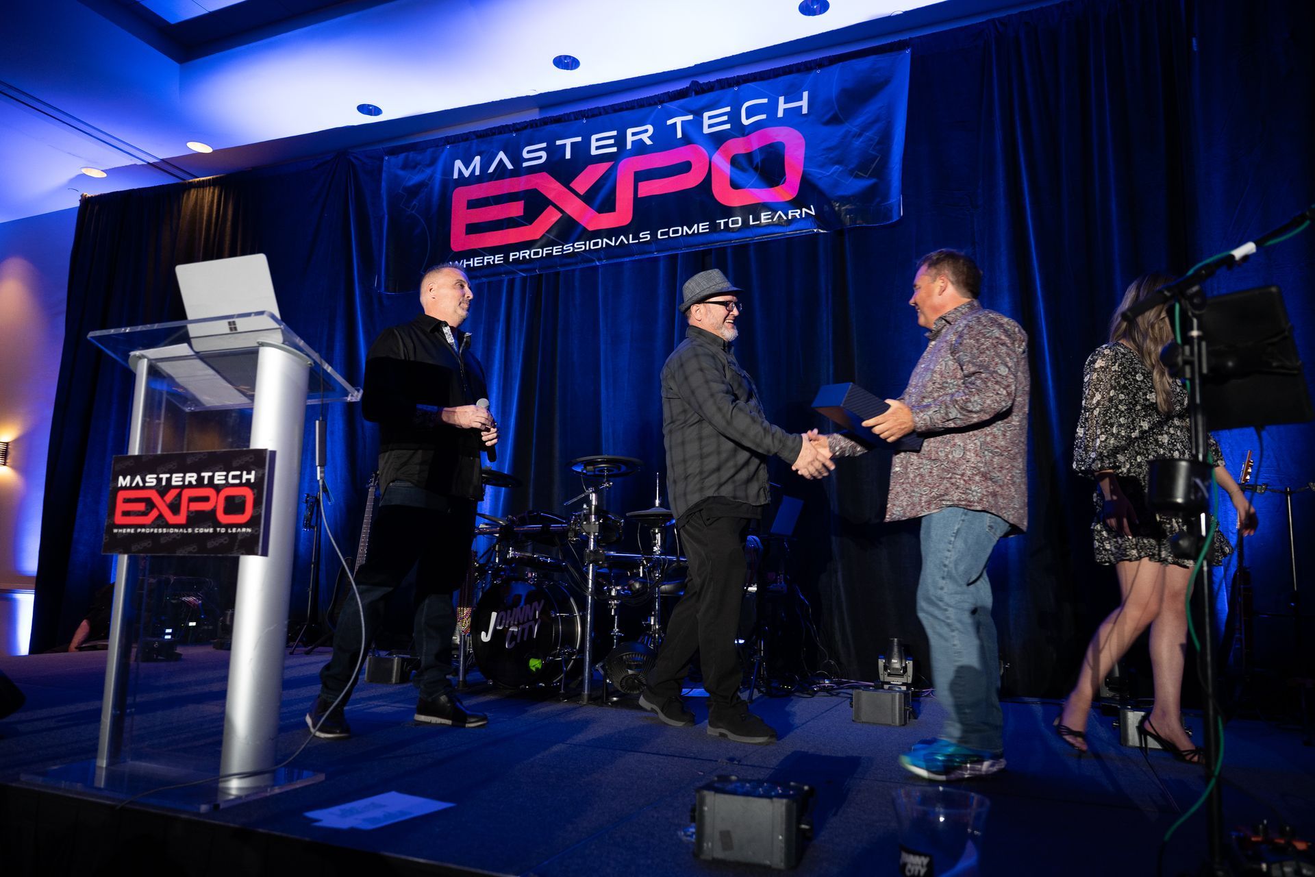 Men shaking hands on stage at Master Tech Expo, banner in background. Other people, drums, and podium are also visible.