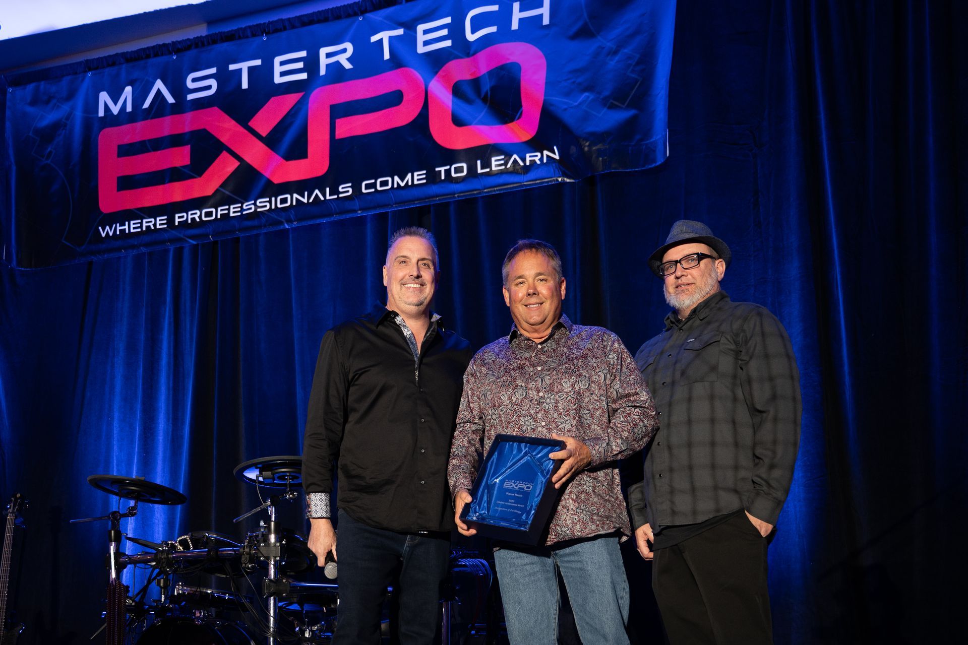 Three men on stage holding an award at the MasterTech Expo. Backdrop banner.