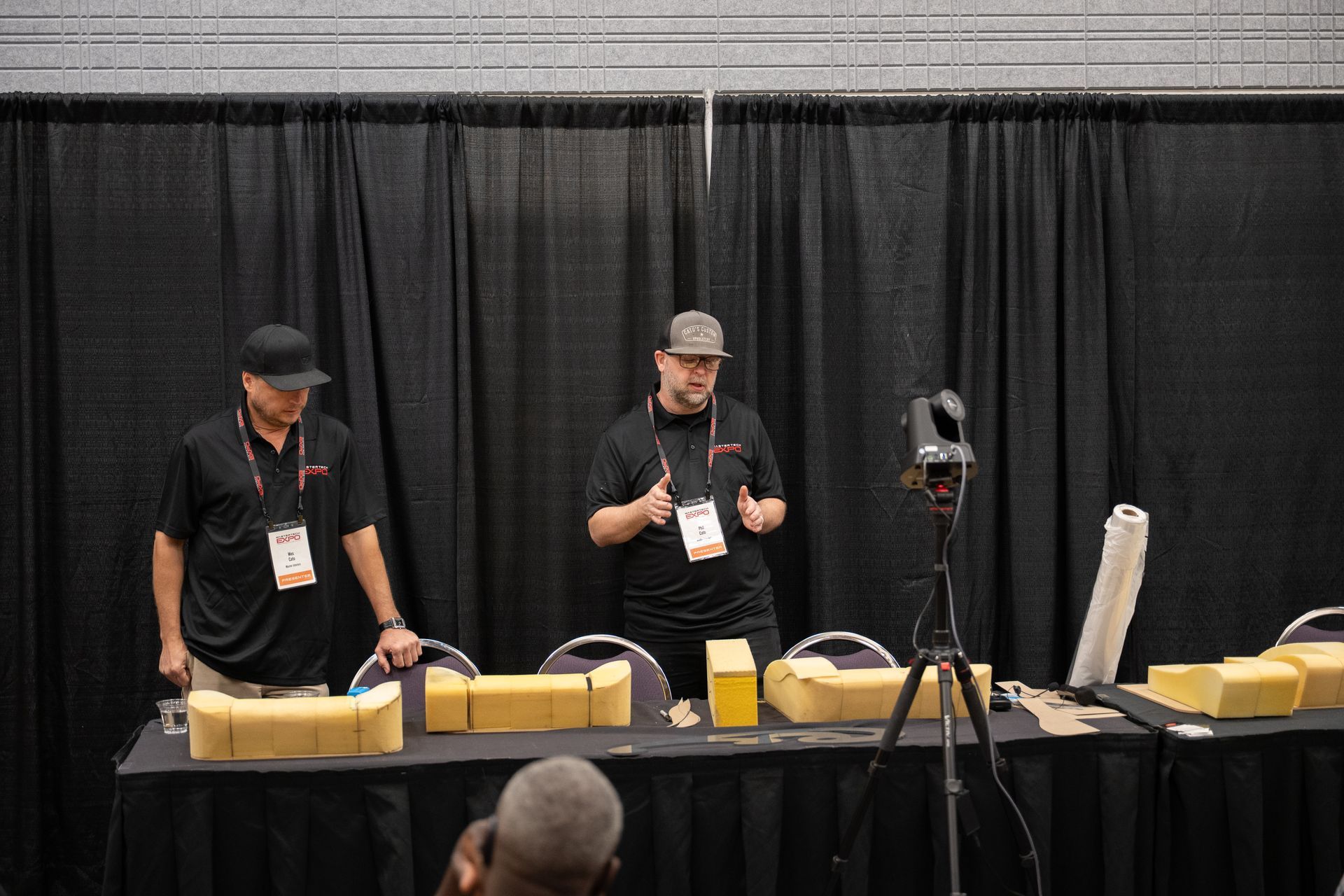 Two men giving a presentation at a table. They stand in front of black curtains and a camera. The table has samples of construction materials.