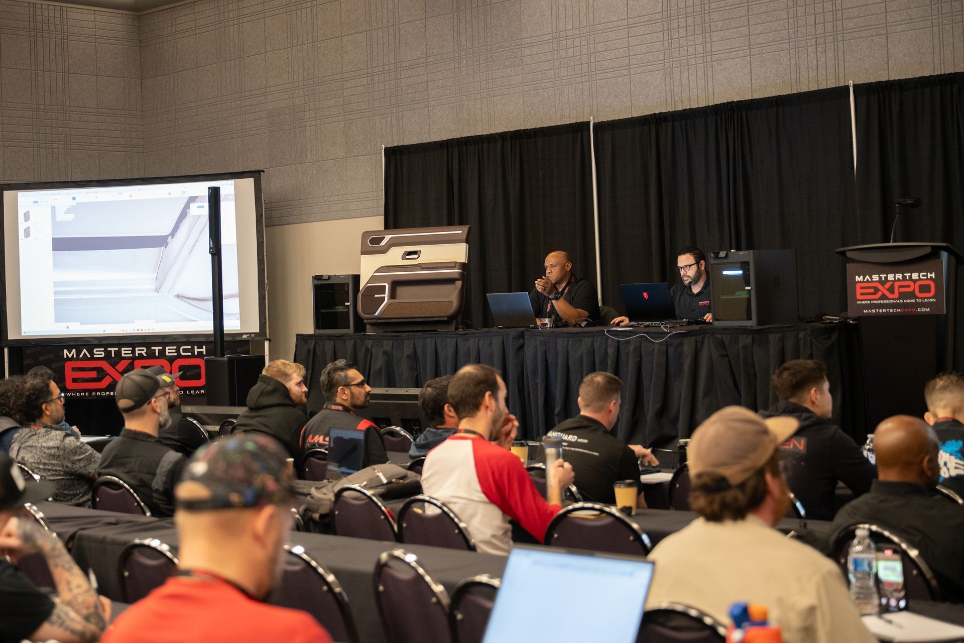 Audience seated at conference; two speakers present at podium with projector screen displaying image.