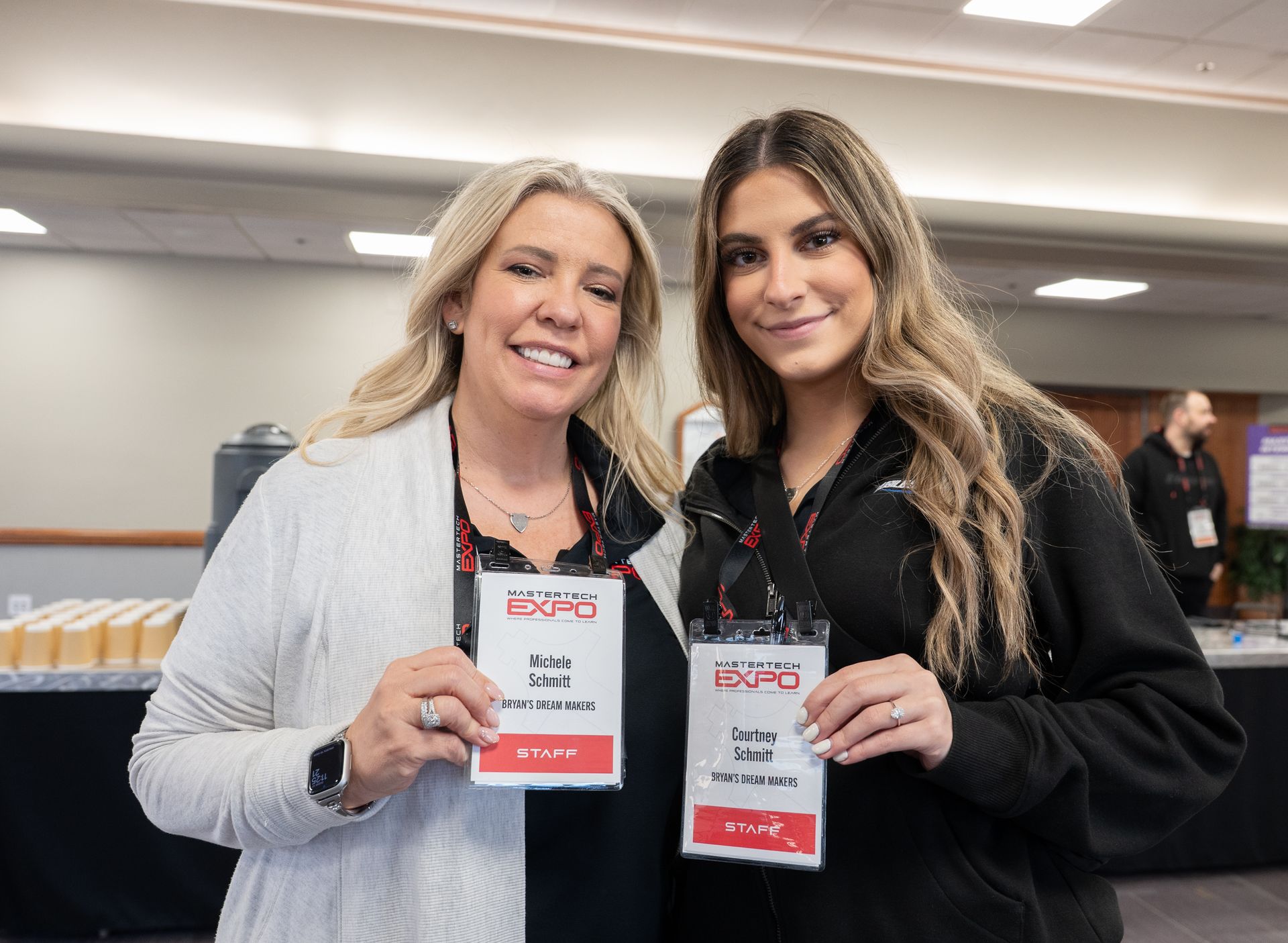 Two women holding event badges, smiling at a conference. One in a gray cardigan, the other in a black jacket.