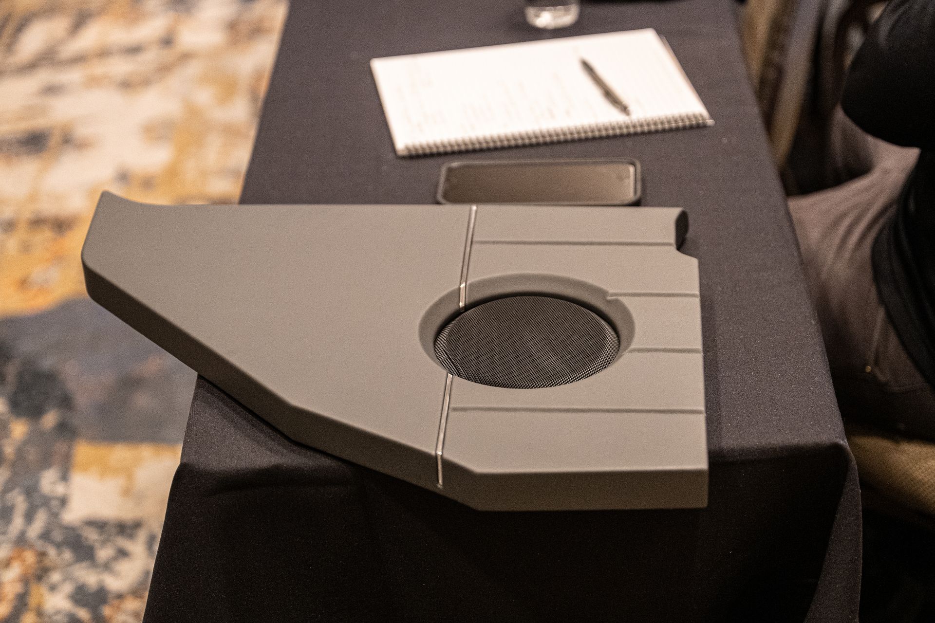 Gray object with a speaker and rectangular design on a black table with a notebook and pen.