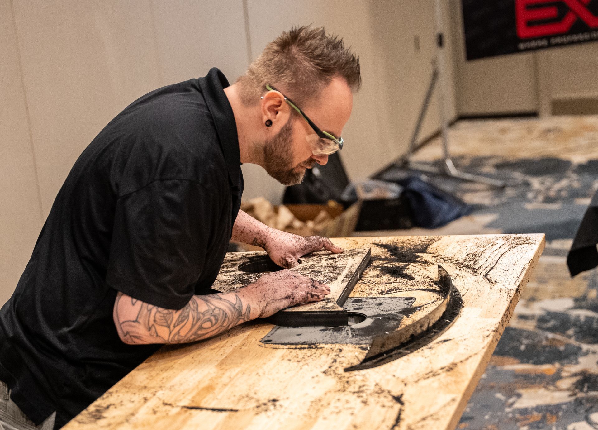Man with tattoos carves a design into a wooden plank, concentrating, in a workshop setting.