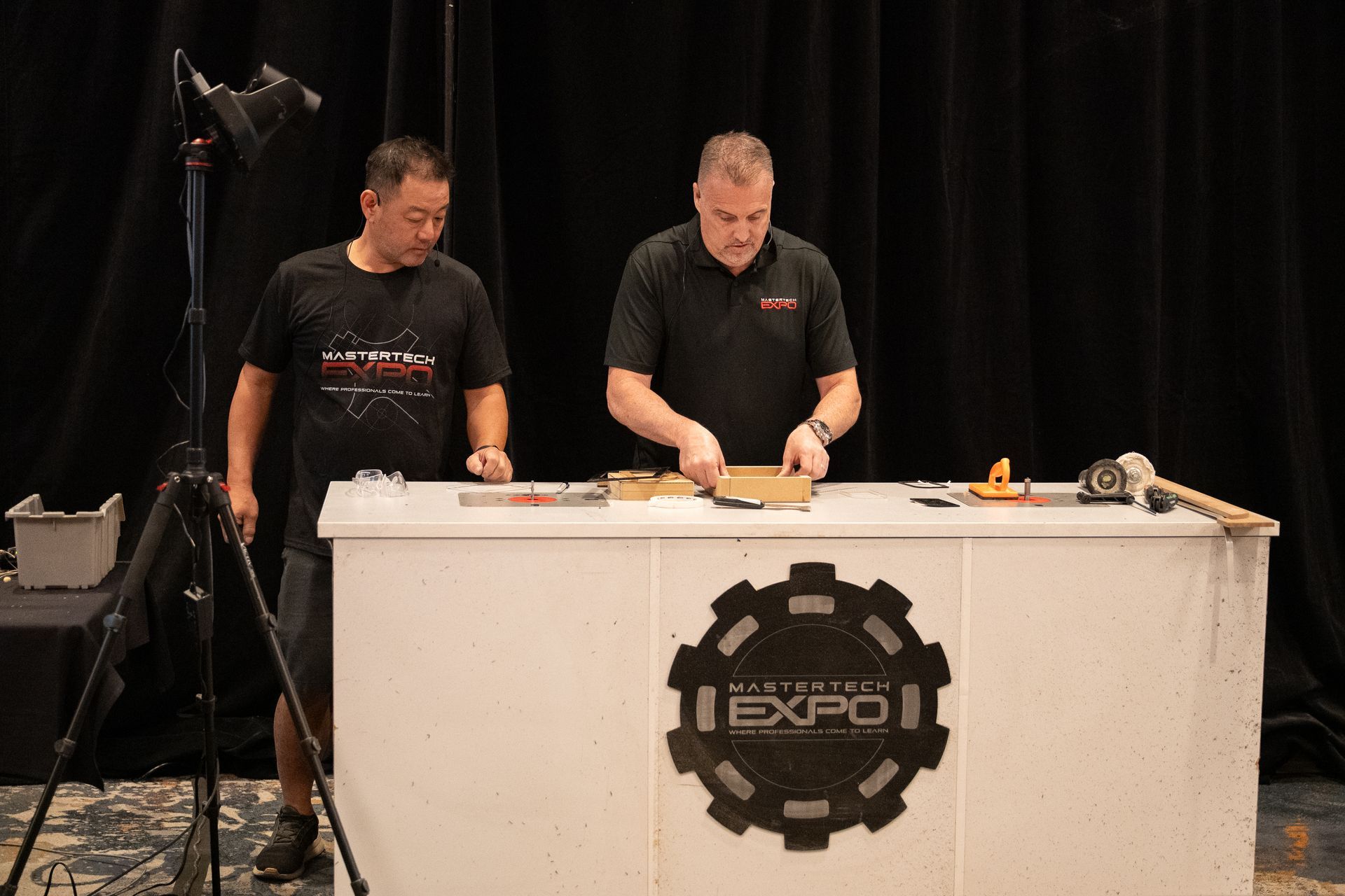 Two men at a booth with tools, assembling something. Backdrop with logo.