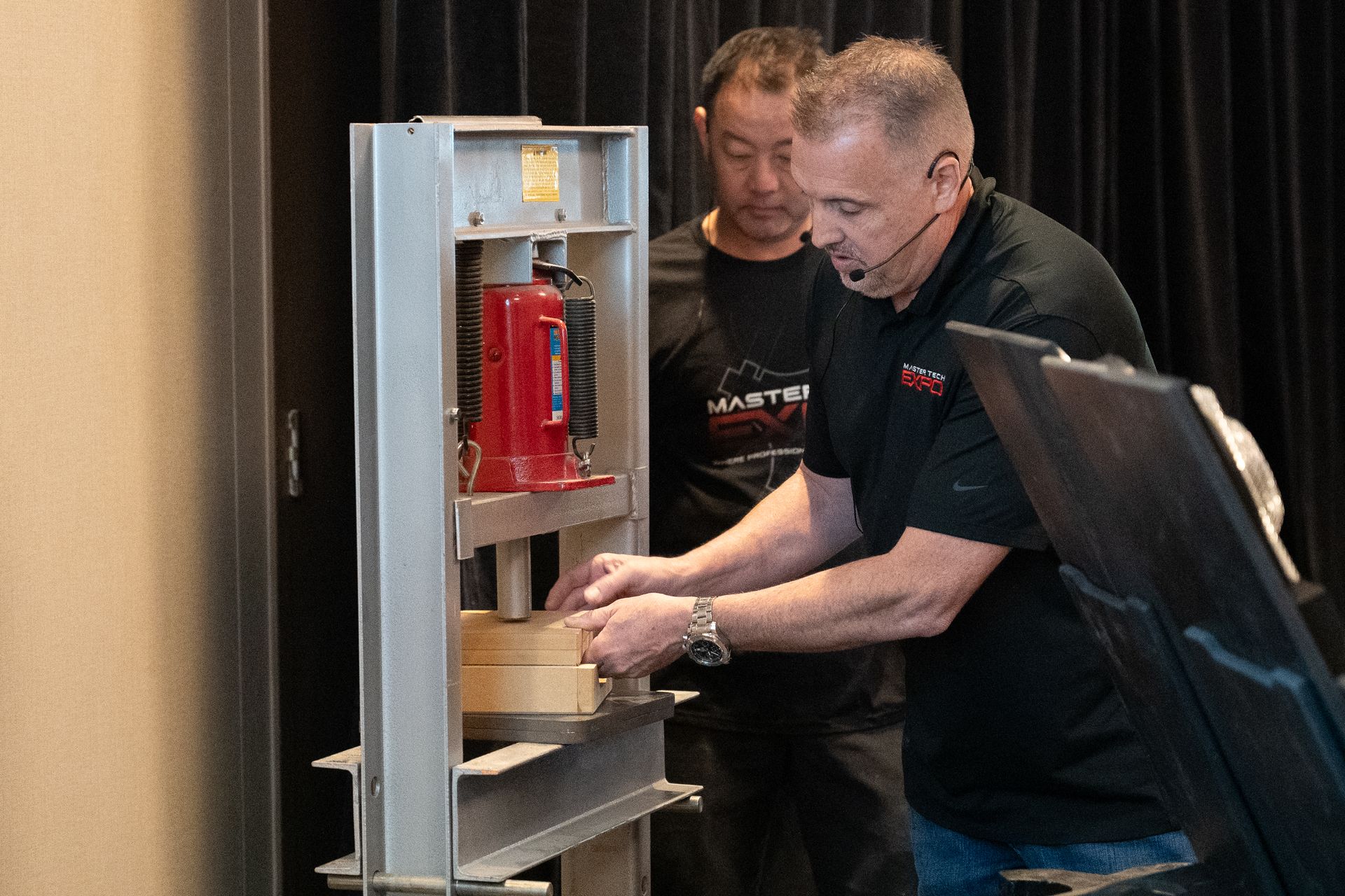 Two men operating a press machine, one pointing, indoors.