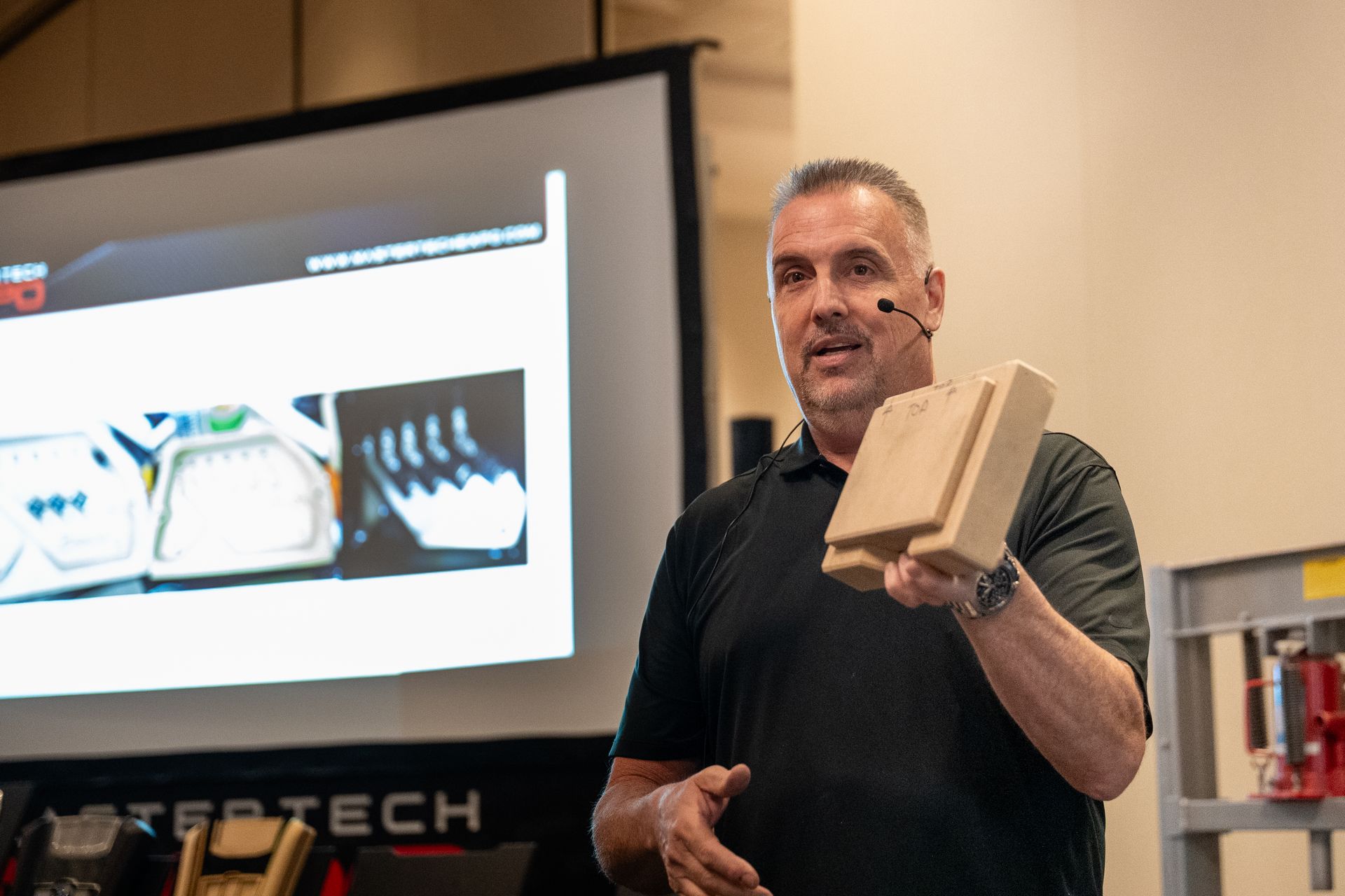 Man presenting a wooden model, standing in front of a projection screen with product images.