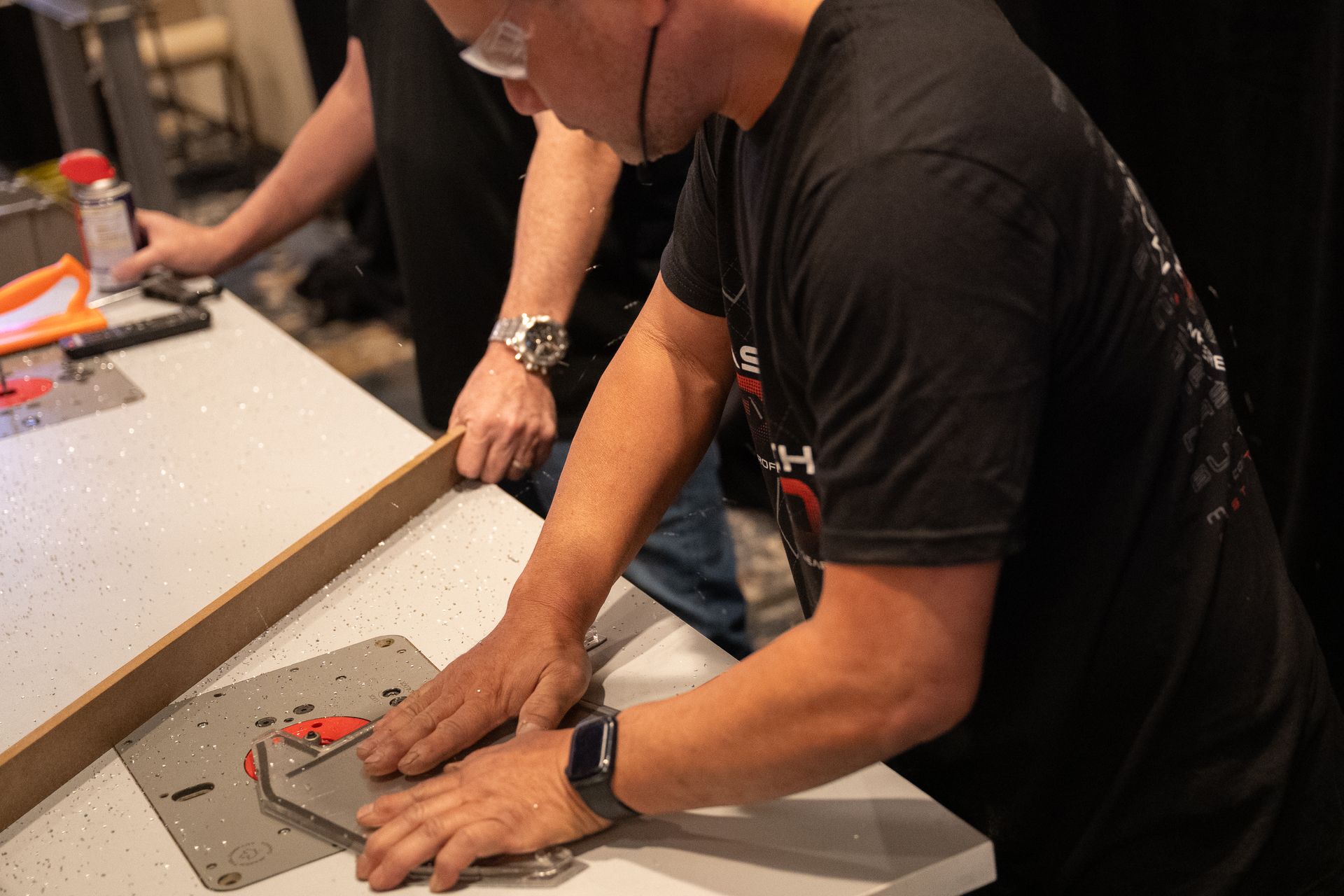Man in black shirt using a power tool on a metal plate at a work table.