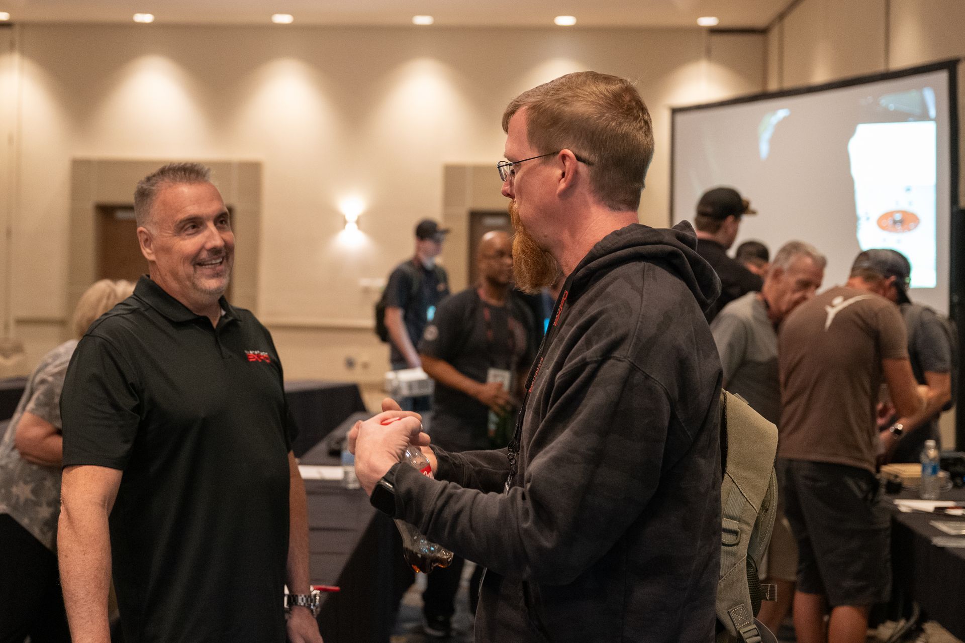 Two men conversing in a conference room. Other people are in the background; a screen is on the wall.