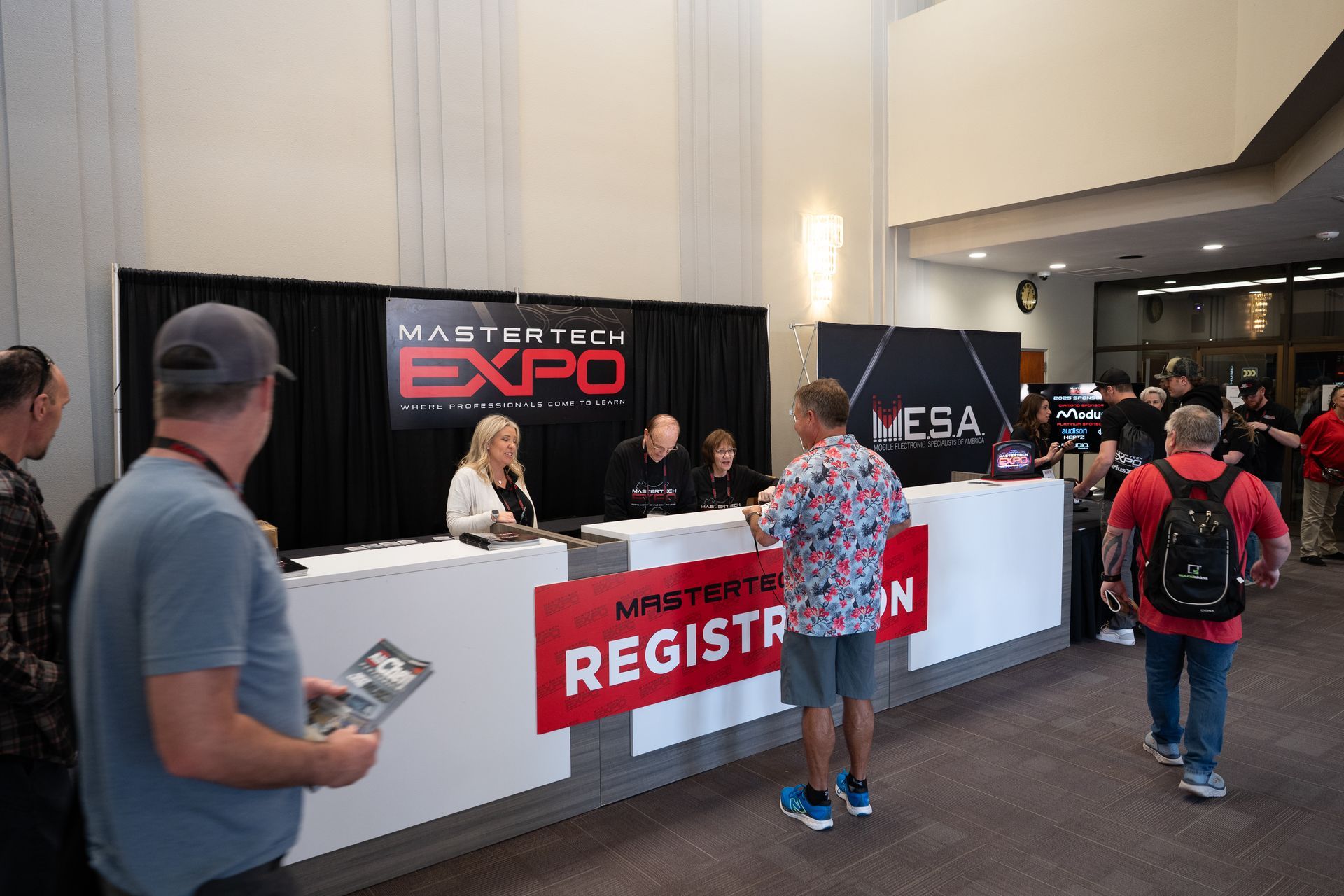 Registration desk at Master Tech Expo. Attendees register, staff seated behind desk. Red 