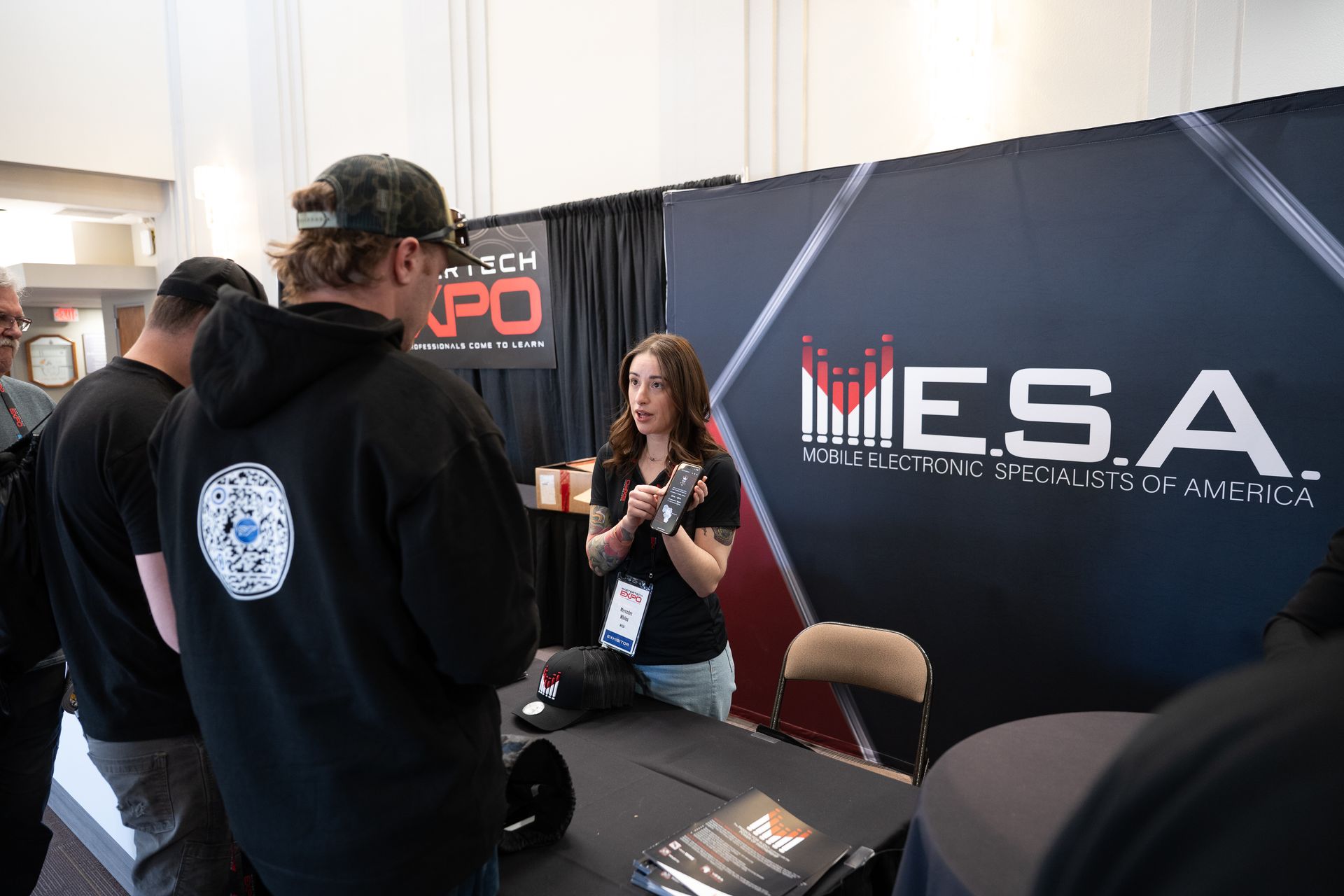 People at a booth for E.S.A. at an expo. A woman holds a product; the background is black with the E.S.A. logo.