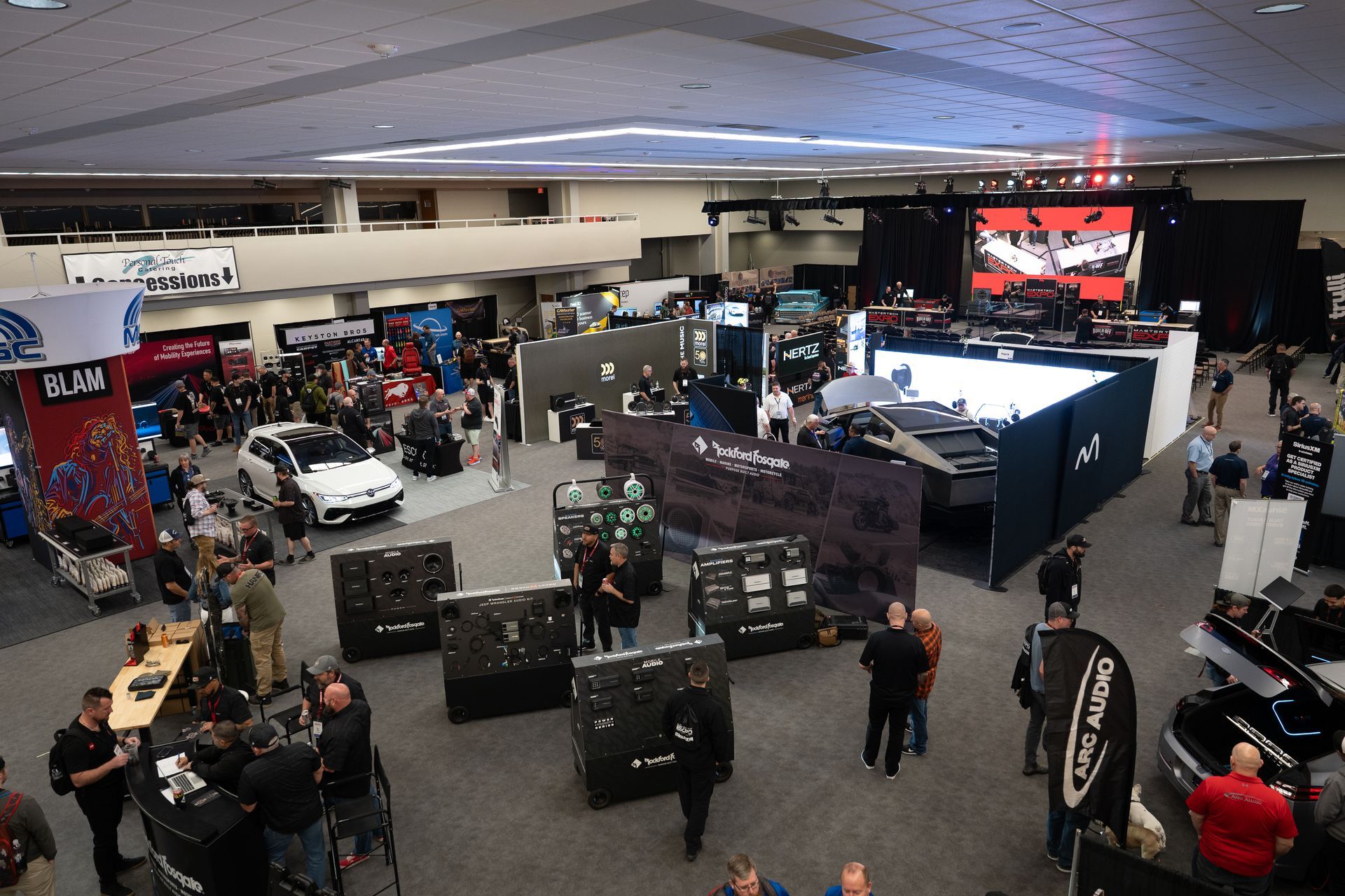 Overhead view of an indoor trade show with vendors, cars, and attendees. Large open space with booths and a stage.