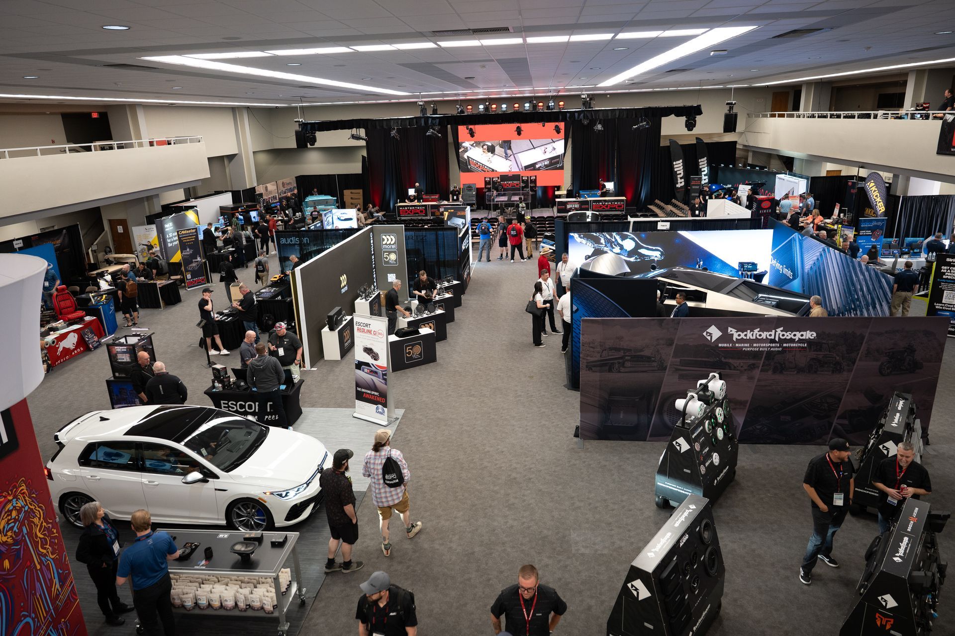 Large convention hall with vendor booths, displays, and people; a white car in foreground.