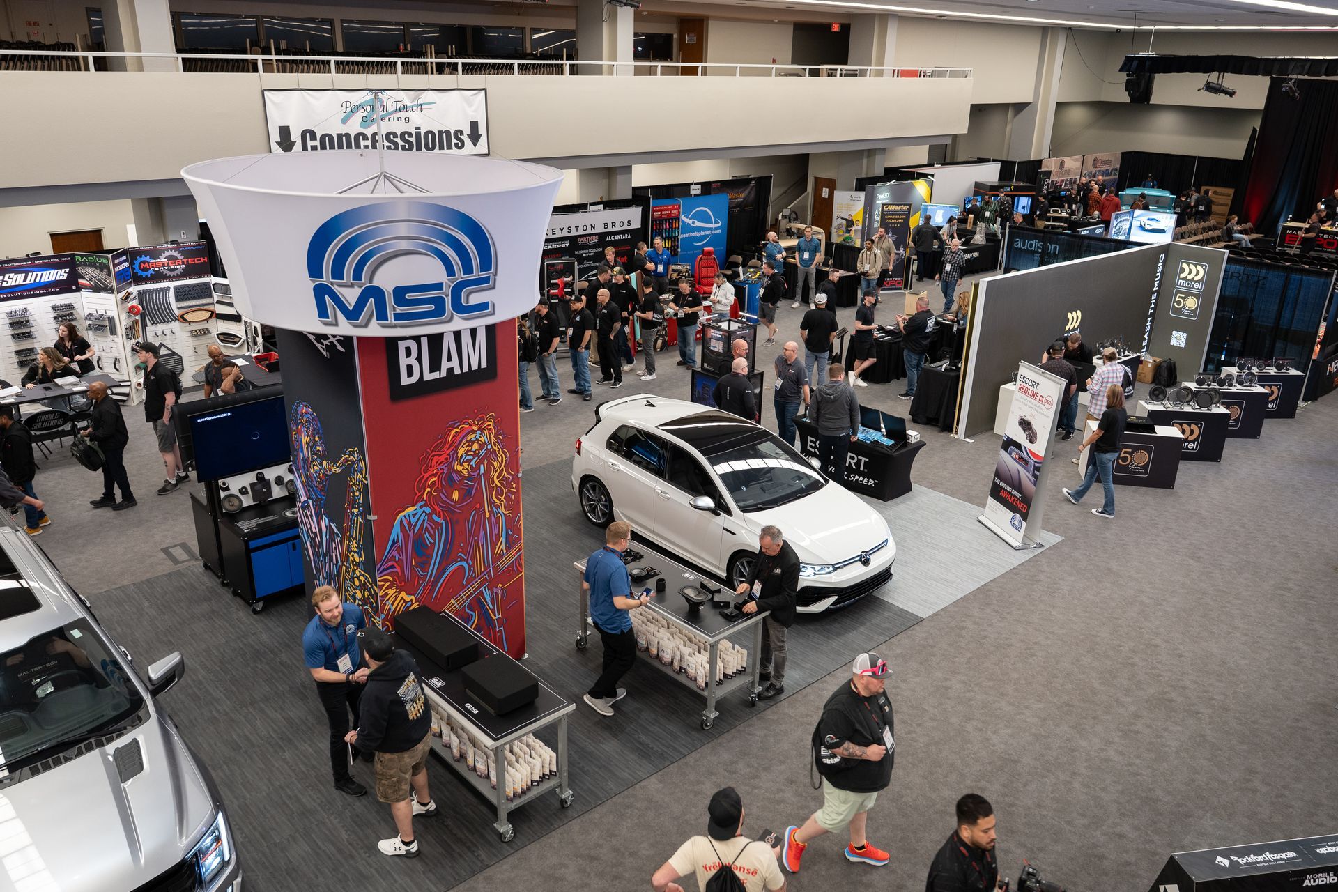 Car audio expo with cars, vendors, and attendees, a large MSC Blam sign overhead.