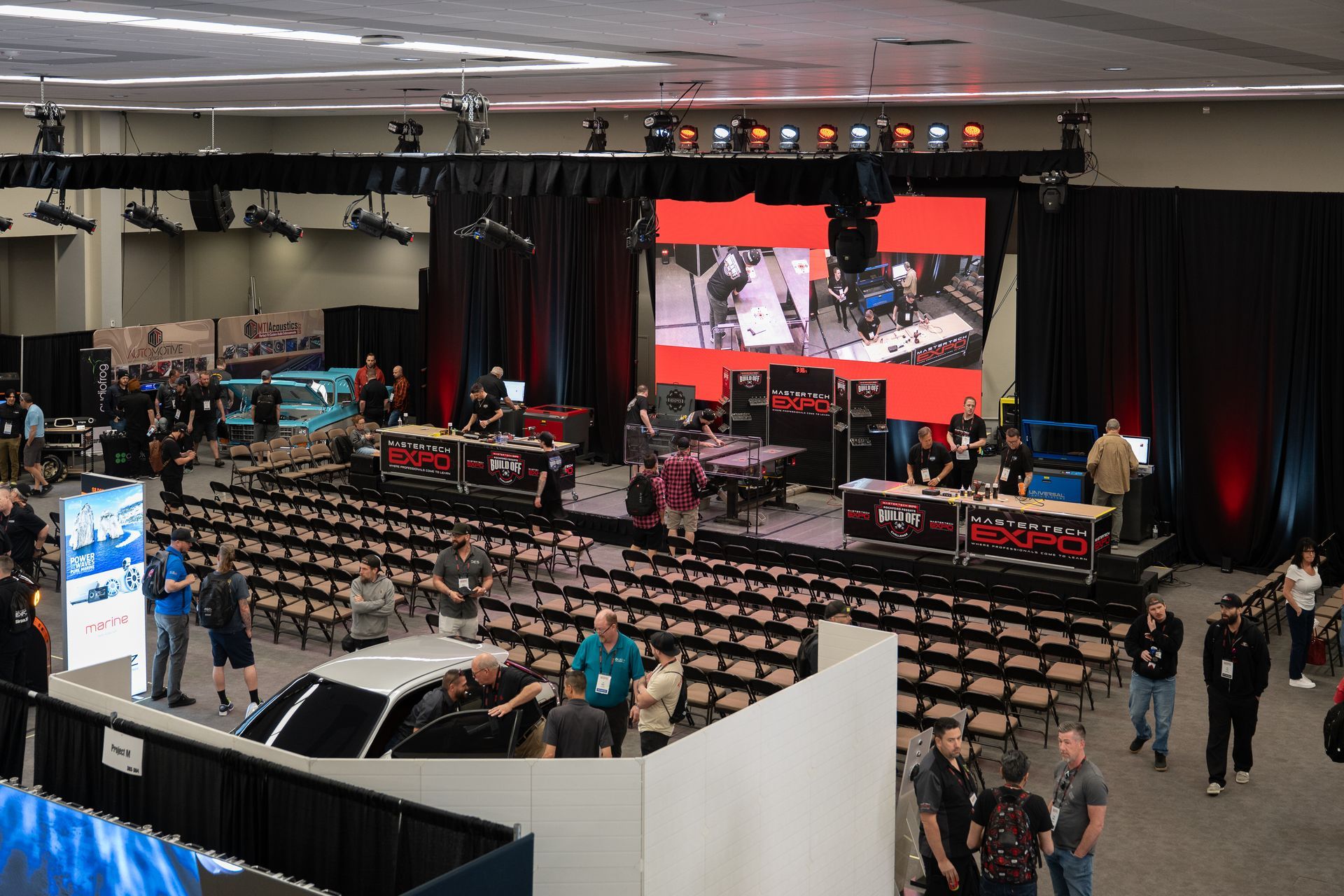 Convention hall with stage, screen displaying images, rows of chairs, and people milling about.