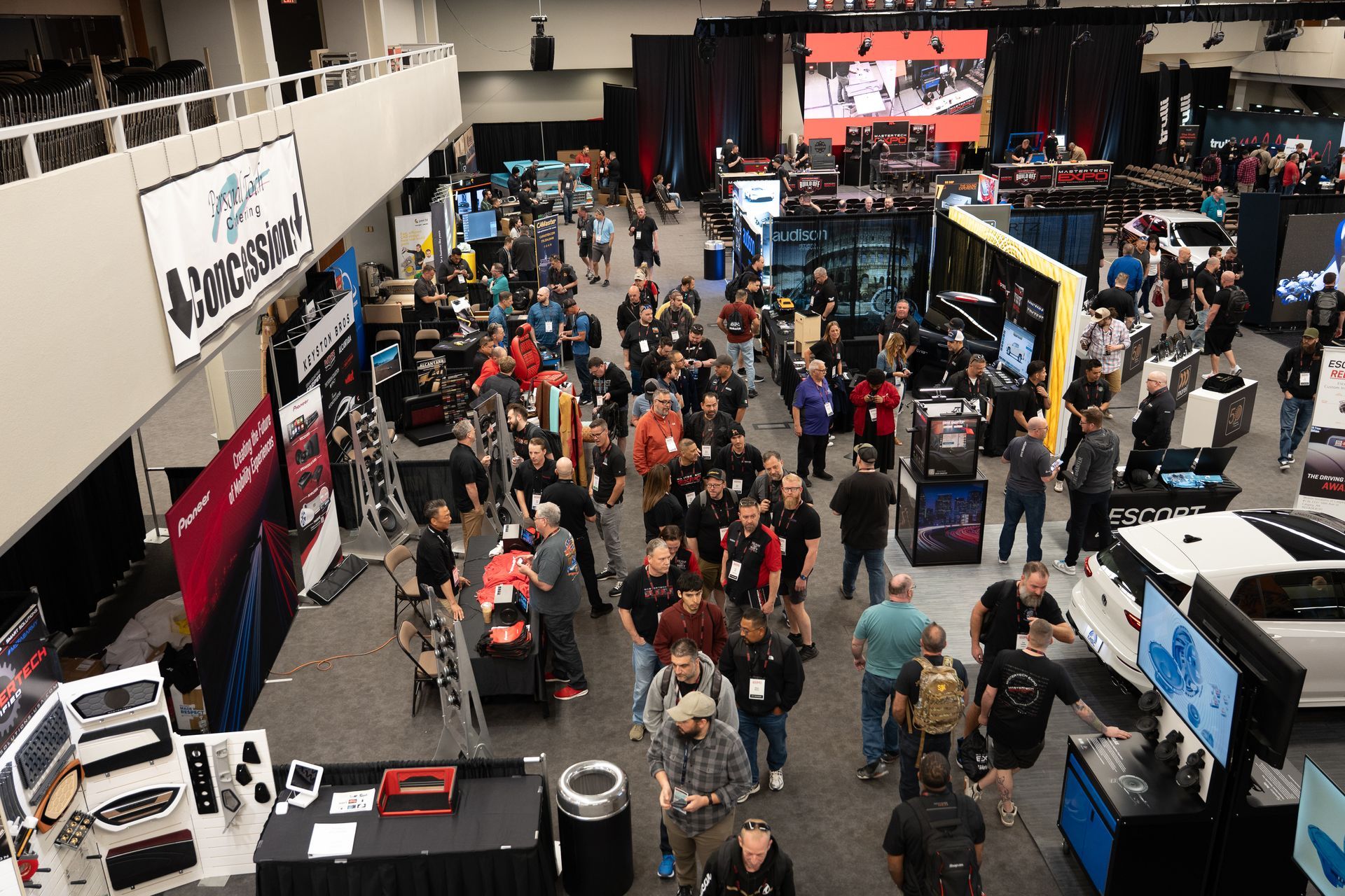 Large convention hall crowded with people viewing exhibits; white cars, screens, banners.