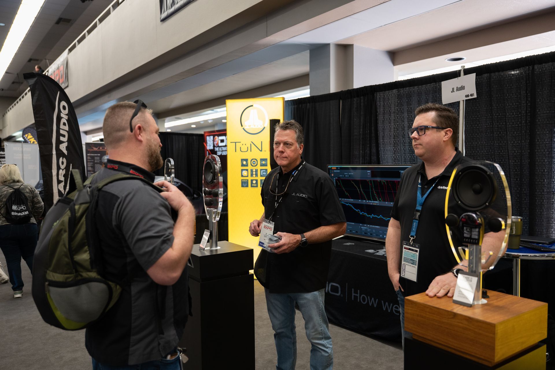 Three men at a booth, discussing audio equipment at a convention. One man holds a speaker.